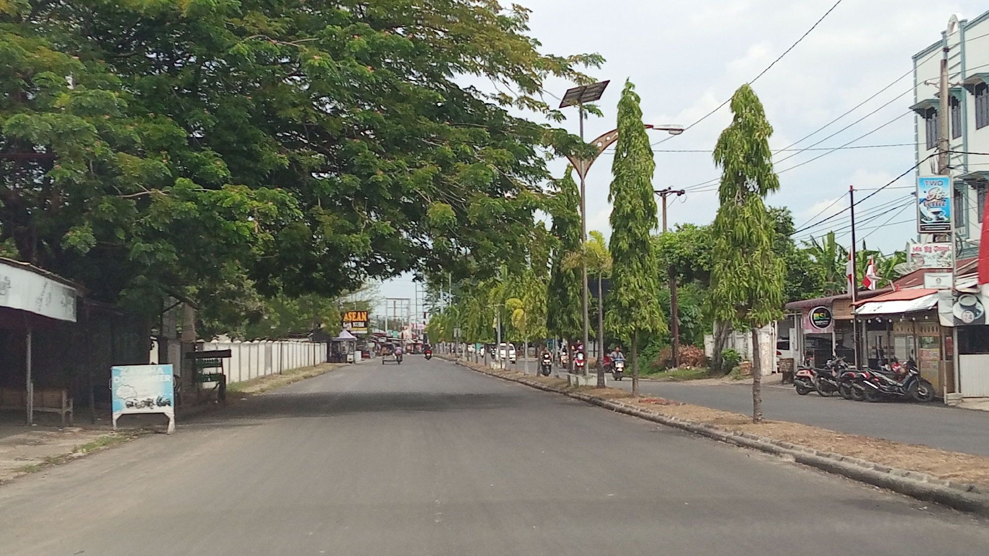 This is an eye-level, wide-angle, full shot of a two-lane asphalt road in Lhokseumawe, Indonesia. The road is lined with trees on both sides, and there are buildings and shops visible on the right side of the road. The sky is overcast.
On the left side of the road, there are large trees with dense green foliage that overhang the road. There is a white fence and a small building with a red roof. A blue sign with white lettering is visible on the left side of the road.
On the right side of the road, there is a row of shops with red roofs. Several motorcycles are parked in front of the shops. Signs are visible on the buildings, including one that says "TWO CUPS Coffee" and another with Indonesian writing. There are also many tall, slender trees planted along the roadside.
Several people are riding motorcycles on the road, and a few are walking. A small cart is being pulled by a person on the left side of the road in the distance. The scene appears to be a busy commercial and residential area.
The overall impression is a bustling street scene in a tropical urban environment.