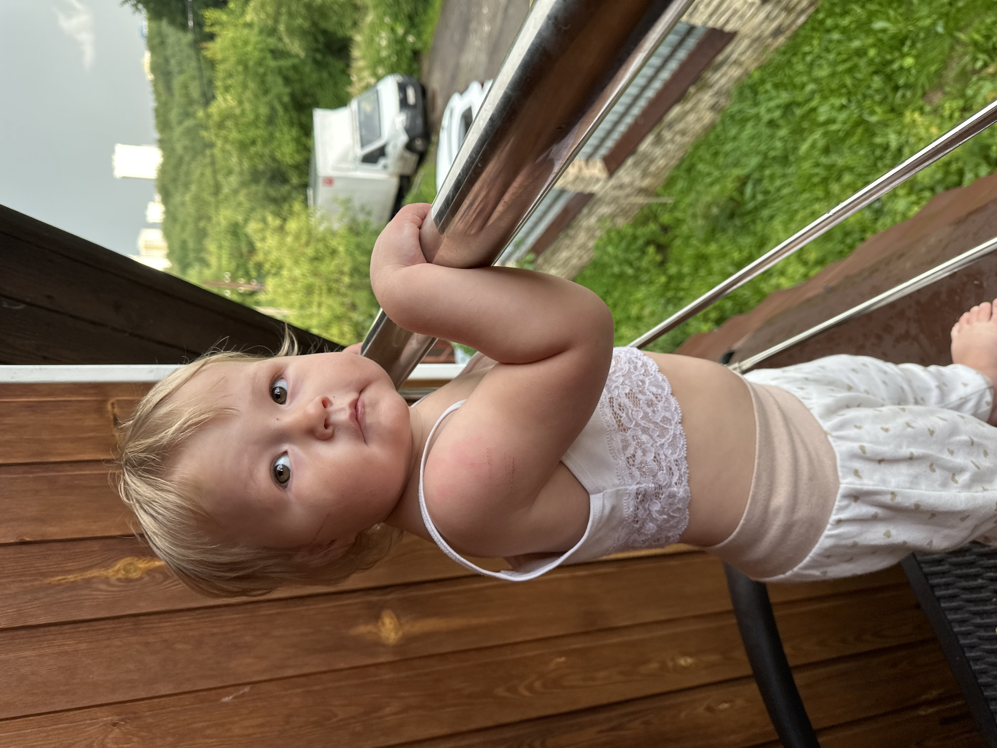A young child with blonde hair, wearing a white lace bralette and white patterned pants, stands on a balcony, leaning on a metal railing. The child is looking upwards and to the right. In the background, there is a wooden wall, lush green trees, a white delivery truck, a parked car, and a few buildings in the distance. The sky appears overcast. The image was taken outdoors in an urban or suburban setting, possibly in Nizhny Novgorod, Russia.