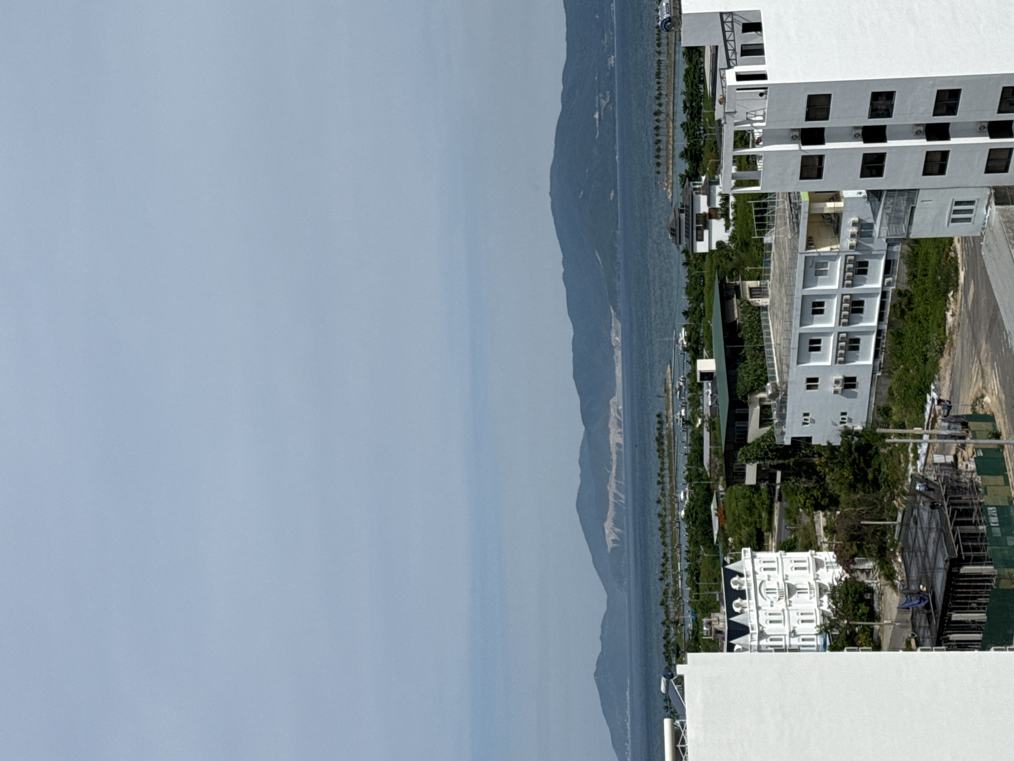 This is a landscape photograph taken in Bac Nha Trang Ward, Vietnam. The image depicts a coastal scene with a body of water on the left, likely the sea, and a mountainous terrain on the right. Several modern buildings with white facades and numerous dark-framed windows are visible along the coastline. One building in particular stands out with its ornate, white architectural details and a series of arched windows. The mountainous area behind the buildings is covered in lush green vegetation. There is a road or pathway running alongside the buildings and leading into the mountainous area. Some construction activity is evident, with scaffolding and tarps visible. The sky is a light blue, suggesting daytime. The overall impression is one of a developed coastal area with residential or commercial buildings situated near a natural mountainous landscape.