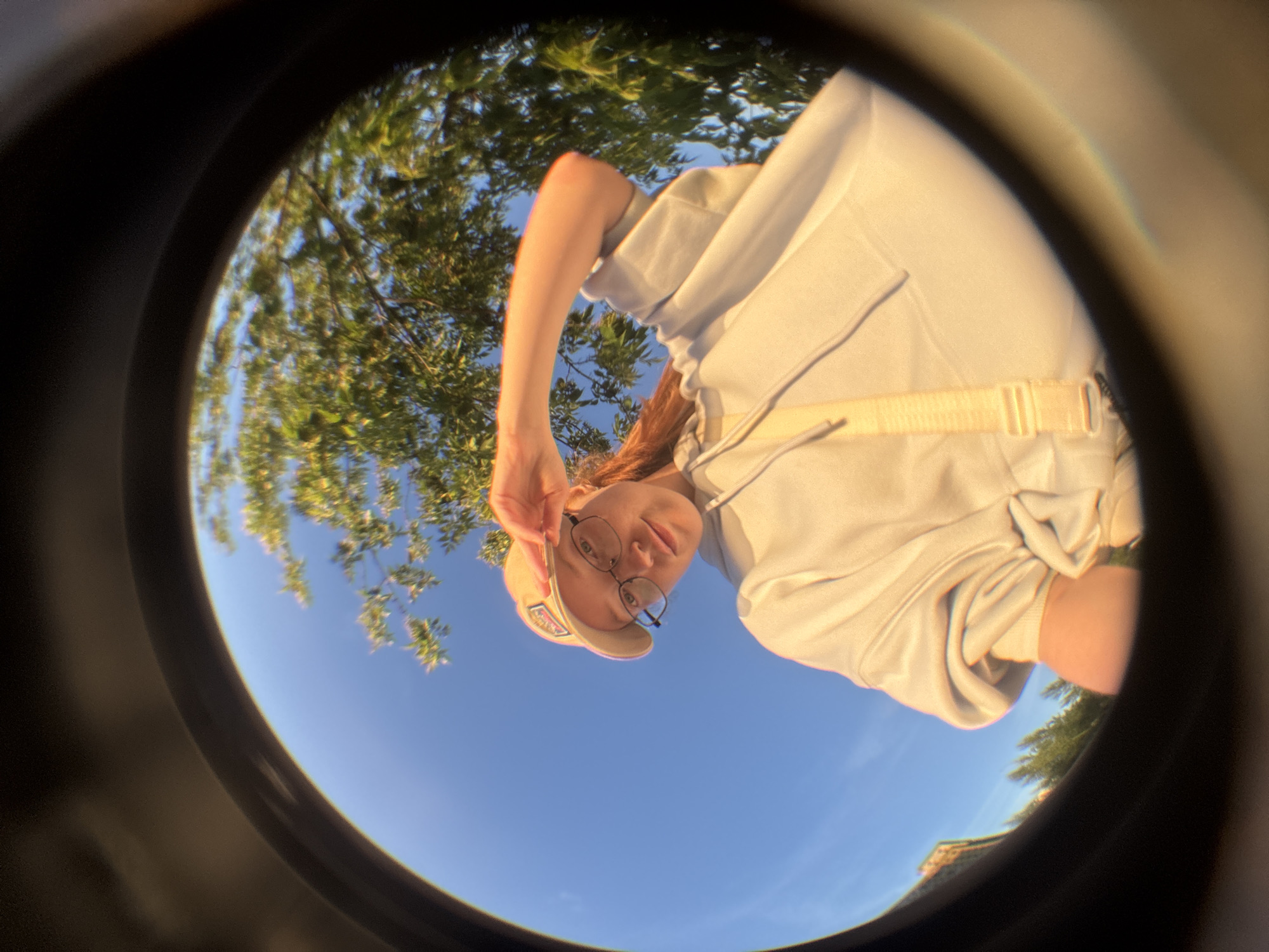 A young woman wearing a light-colored hoodie, a baseball cap, and glasses is captured in a fisheye lens perspective. She is outdoors, with a clear blue sky and lush green trees forming the background. The woman is looking up and to her right, with her right arm raised and hand resting on the brim of her cap. A light-colored strap is visible across her chest. The image appears to be a selfie taken from a low angle. The location is not identifiable as Saint Petersburg, Russia.