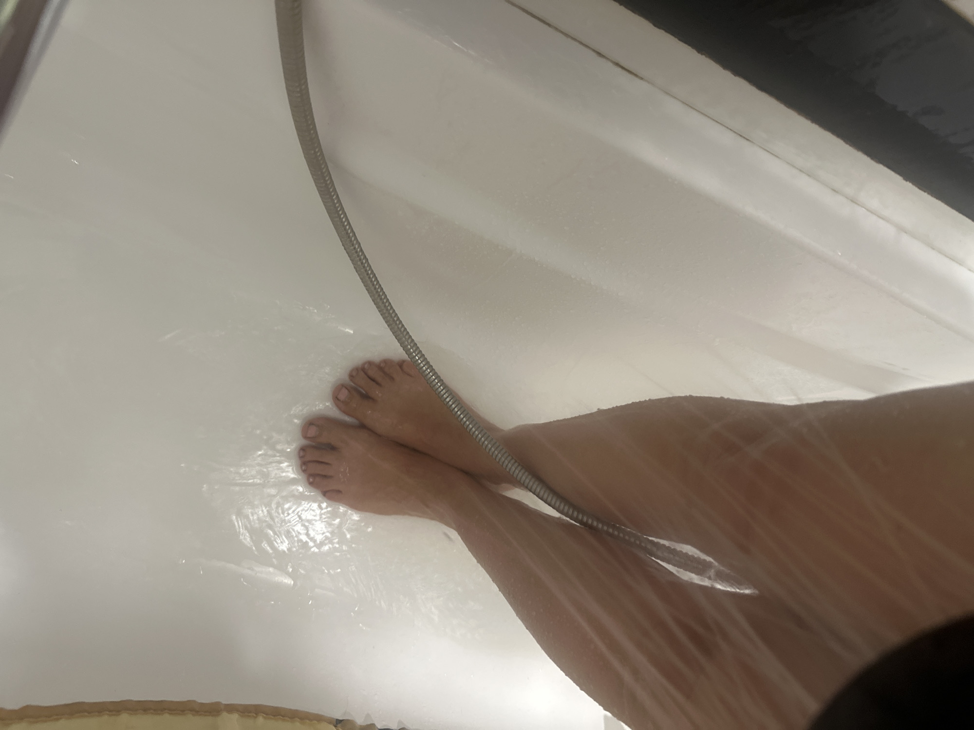 The image shows a person's legs and feet in a shower. Water is visible, creating a blurry effect on the skin. A shower hose is present on the right side of the frame. The location is identified as Novoaltaysk, Russia.