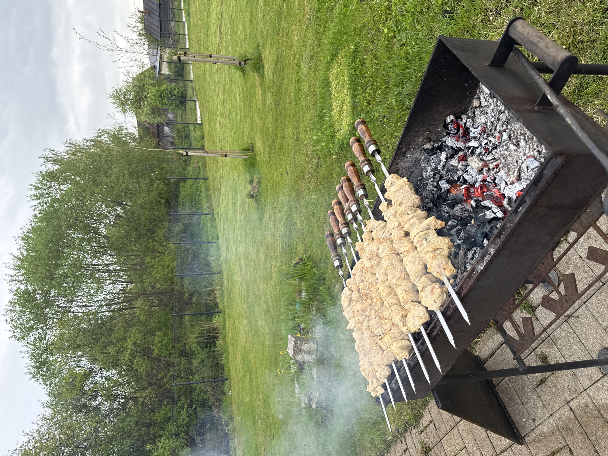 Outdoor scene in Rezekne, Latvia. A metal barbecue grill sits on a brick-paved area, with skewers of meat (likely shashlik) cooking over smoldering charcoal. The grill is black with a visible handle. The skewers have wooden handles and are arranged along the grill. Smoke rises from the grill. In the background, there is a grassy lawn with trees and shrubs, and a wooden fence can be seen in the distance. The sky is overcast. The photo is taken at an angle, showing the grill and the skewers as the primary focus.