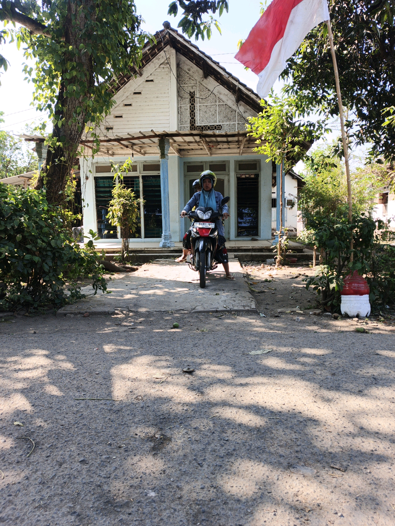 A person wearing a light blue headscarf and a green helmet is seated on a motorcycle parked on a paved surface in front of a white house with a red and white flag flying above it. The house has a porch supported by blue pillars and a pitched roof. Lush green trees and bushes surround the house and the motorcycle. The ground in front of the house is covered in asphalt with patches of dirt and scattered green leaves. A large tree with visible vines stands to the left of the house, its branches partially obscuring the building. To the right of the house, a white plastic bottle with a red top is placed near the bushes. The scene appears to be set in a rural or suburban area of Modongan, Indonesia, given the presence of the Indonesian flag and the architectural style of the house.