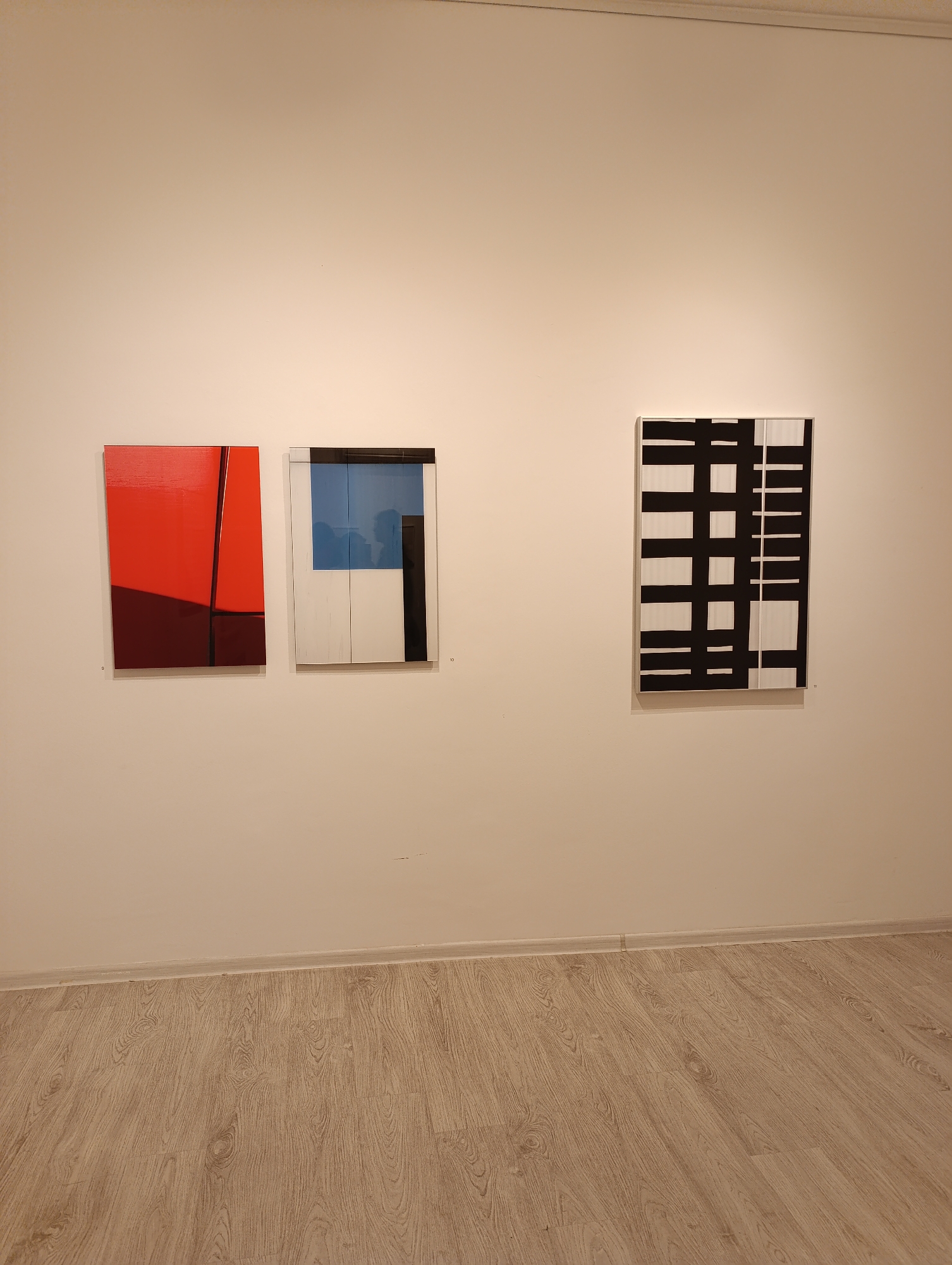 The image depicts an interior scene, most likely an art gallery. There are three abstract paintings hanging on a white wall. The painting on the left is primarily red, with a dark red section and a black diagonal line. The middle painting features blue, white, and black sections within a silver frame. The painting on the right is black and white and appears to have a grid pattern. The floor is made of light brown wood planks. No people are visible. No information about the location in Sofia, Bulgaria, can be discerned from the image.