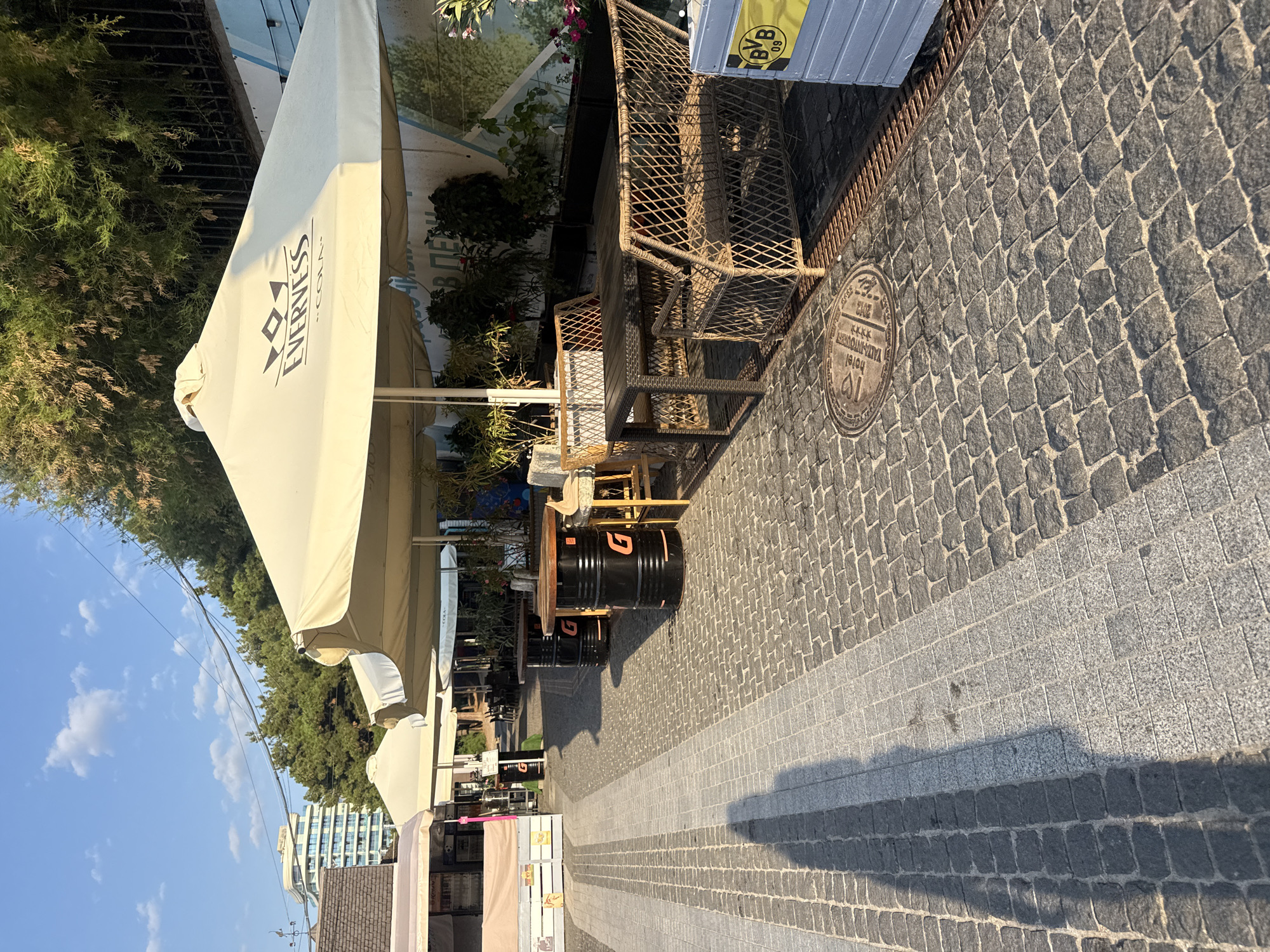 The image depicts an outdoor seating area of a cafe or restaurant on a cobblestone walkway in Yalta, Ukraine. Several large, cream-colored umbrellas, one branded with "EVERNESS", provide shade over tables and seating. Some tables are made from repurposed black oil barrels with wooden tops. The seating includes wicker chairs and benches. In the background, lush green trees and a multi-story building are visible against a clear blue sky. A shadow of a person is cast across the walkway in the foreground. A manhole cover is visible on the cobblestones. A grey container with a yellow "BVB 09" logo is present on the right side of the image.