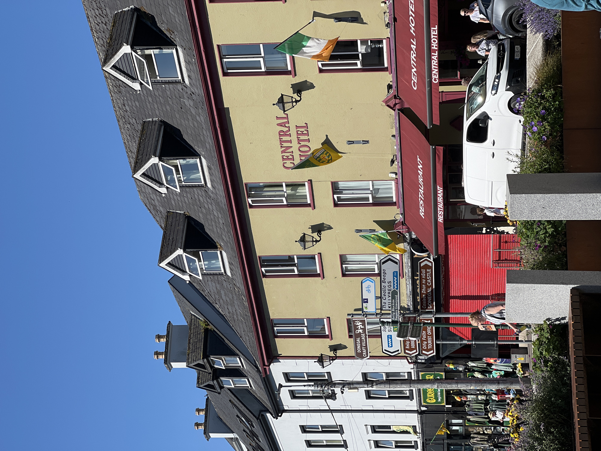The image depicts a street scene in Donegal Town, Ireland, focusing on the Central Hotel and surrounding buildings. The Central Hotel, painted a pale yellow with a maroon awning, dominates the right side of the frame. An Irish flag and a green and yellow flag are displayed on the hotel's facade. Two signs for "RESTAURANT" and "CENTRAL HOTEL" are visible under the awning. To the left of the hotel, a white van is parked in front of the building.
Further to the left, a white building with black trim stands adjacent to the hotel. A street lamp and a pole with multiple directional signs are positioned in front of these buildings. These signs point to various locations within Donegal Town and its vicinity, including "Donegal Craft Village," "The Castle Begga," "Killybegs," "Ruins of Donegal Abbey," "Oifig Fáilte Tourist Office," and "Caisleán Dhún na nGall Donegal Castle."
On the far left of the image, a shopfront displays an array of clothing and merchandise. A woman is walking past the shop, holding a bottle, and a few other individuals are seen near the van. The foreground features a planter with purple flowers and a bench. The sky is clear and blue, indicating a sunny day.