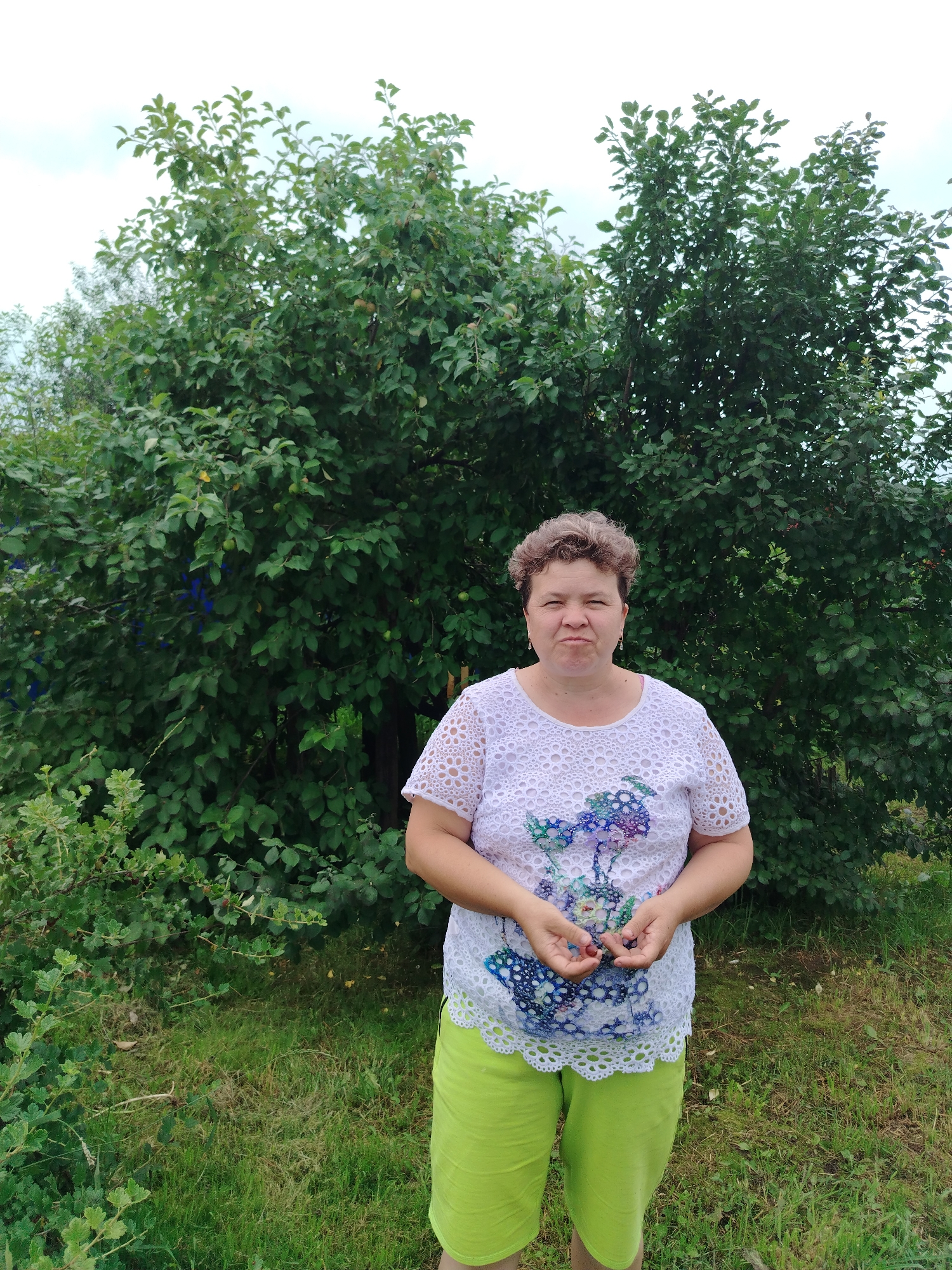 A middle-aged woman with short, curly brown hair is standing in front of lush green trees and bushes. She is wearing a white lace top with a floral design and bright green shorts. Her arms are bent at the elbows and her hands are clasped in front of her, holding a few dark berries. She is looking directly at the camera with a slight pout. The background consists of dense foliage and a slightly overcast sky. The setting appears to be a garden or a park. The image was taken in the city of Gzhatsk (now Gagarin), Russia.