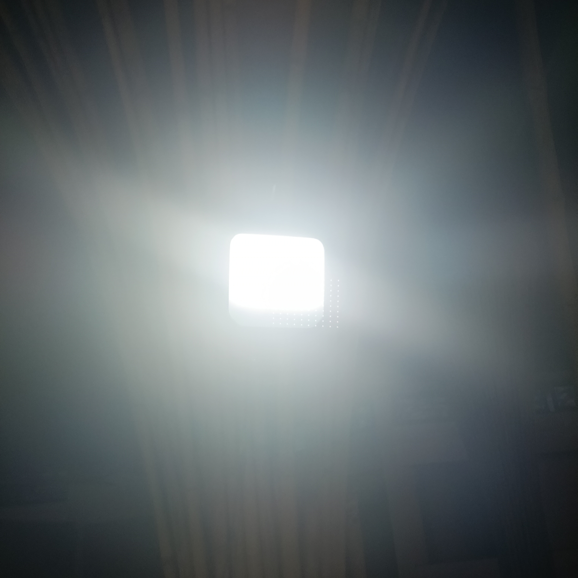 The image shows a bright, square light fixture in a dark environment. The light is the most prominent feature, emitting a strong glow that obscures the surrounding details. Faint vertical lines are visible in the background, suggesting a textured wall or curtains. There is no indication of the image being taken in Panaji, India, or involving any specific people, subjects, objects, or interactions. The image appears to be a close-up shot of a light source.