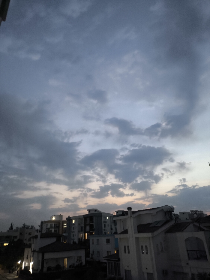 The scene depicts an urban residential area in Nicosia, Northern Cyprus / Cyprus, during dusk or early evening. The sky, occupying approximately two-thirds of the frame, is overcast with dense, dark grey and blue-grey cloud formations, transitioning to a subtle, pale orange hue at the horizon line. Below, a cluster of residential buildings is visible. These comprise multi-story modern apartment blocks with some illuminated windows, and detached or semi-detached houses characterized by white facades and dark-tiled roofs. Several rooftops feature cylindrical solar water heaters. Sparse dark tree foliage is present between structures. No individuals are depicted, nor are any specific actions or interactions discernible.