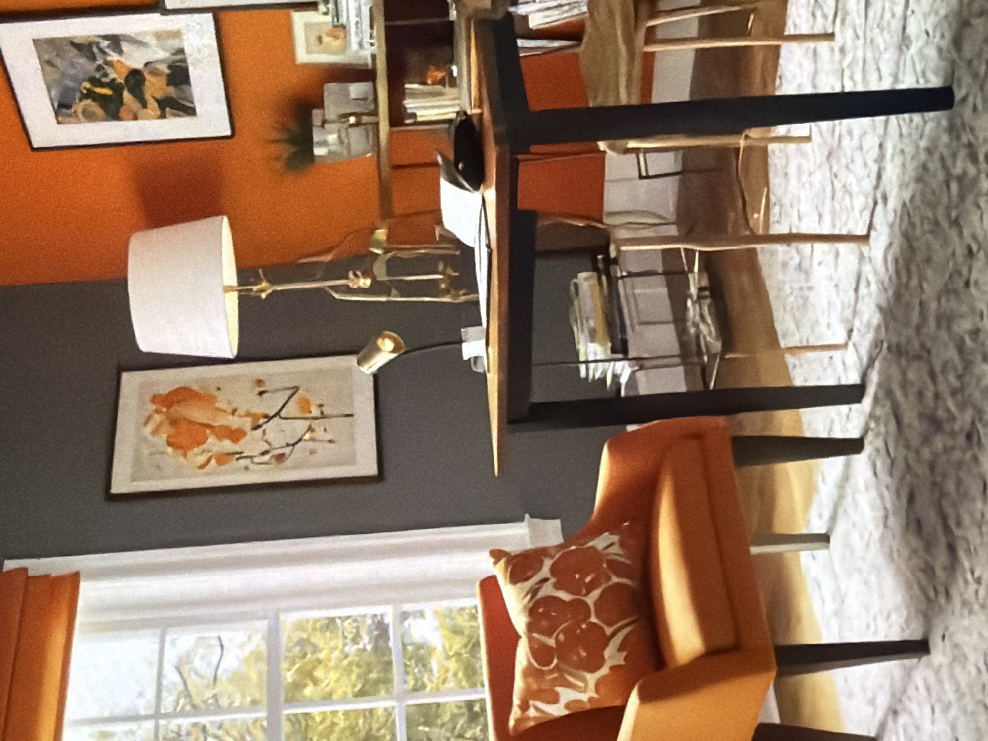 The image depicts a home office or living space with a vibrant color scheme. In the foreground, an orange armchair with a white and orange floral patterned pillow is visible. The armchair is placed on a light-colored rug. Behind the armchair, a wooden table with dark legs is positioned. On the table, there is a lamp with a white shade, a book, and other desk items. To the right of the table, a shelf unit holds books and decorative items. The background features an orange accent wall and a dark gray wall. A framed artwork with orange abstract elements hangs on the dark gray wall, and another framed artwork with a colorful abstract design is on the orange wall. A window with white blinds is on the left side of the image, allowing natural light to enter the space. There are no people in the image.