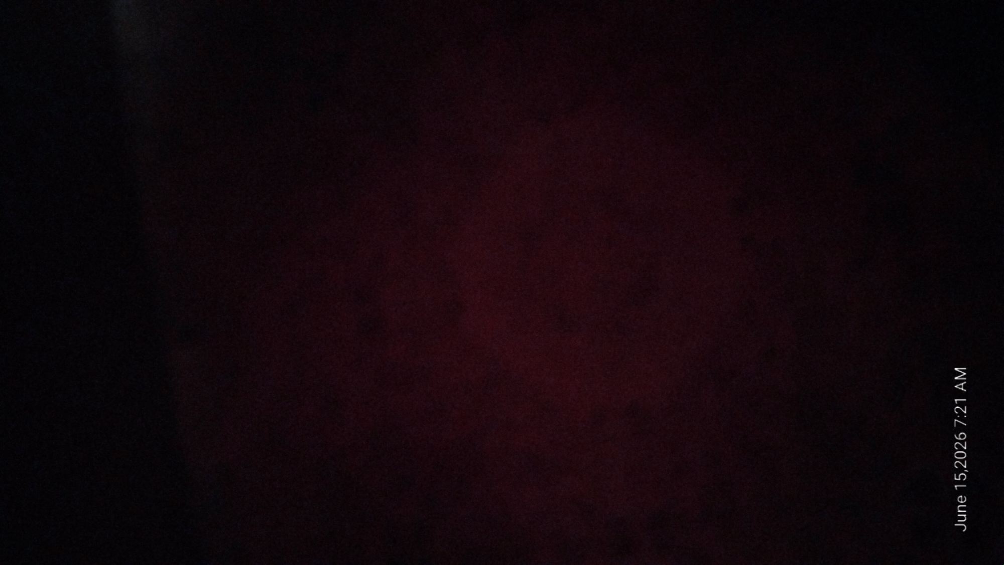 The image is a low-light photograph showing a dark, reddish-brown textured surface, possibly fabric or a wall. In the bottom left corner, white text reads "June 15, 2026 7:21 AM". There are no people, subjects, objects, actions, or interactions clearly visible in the scene. The environment is not discernible due to the darkness. The image does not provide any information about the location in the city of Howayi, Nigeria.