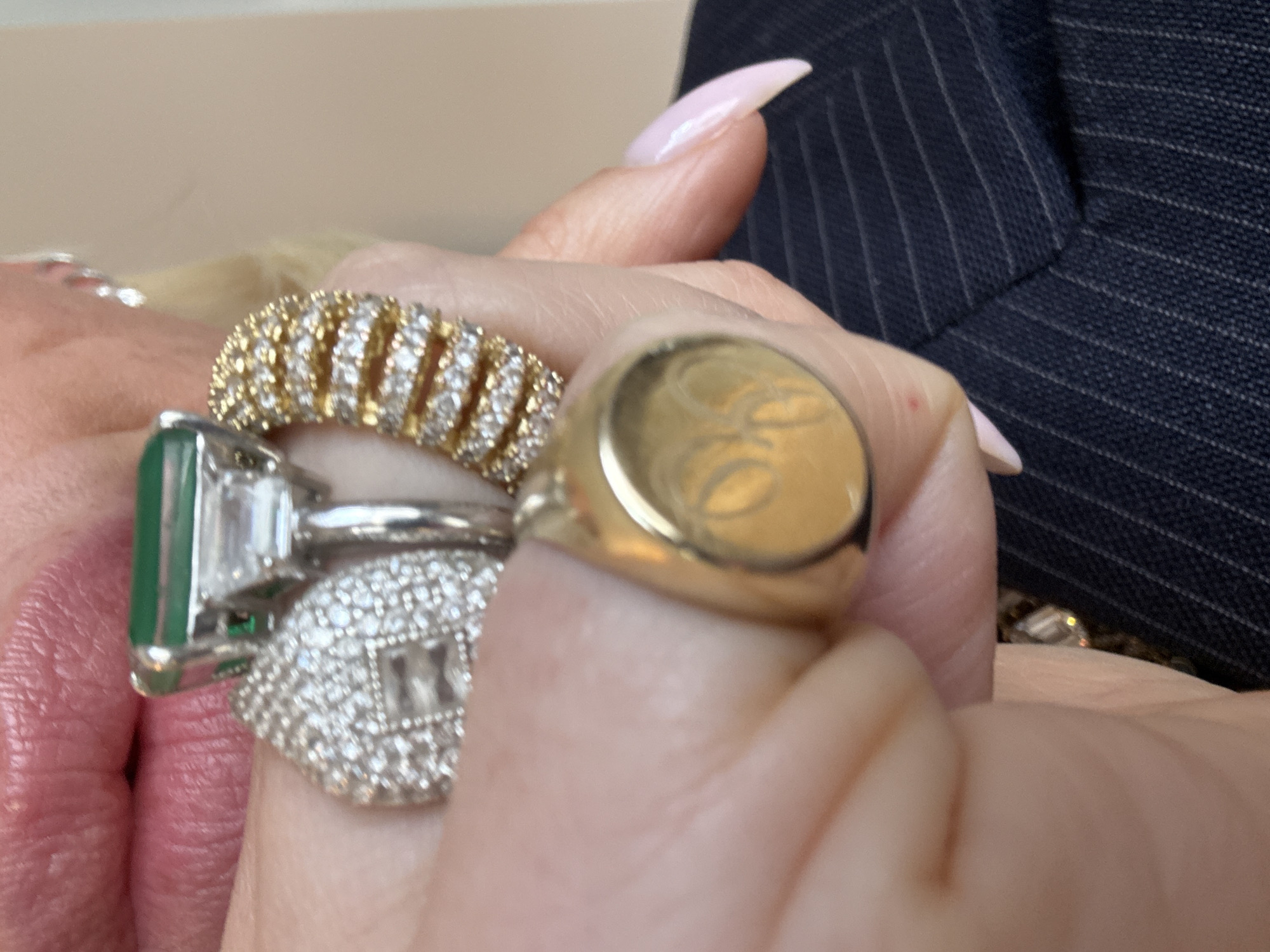 A close-up image focuses on a person's left hand and lower facial area, specifically the lips. The hand displays four prominent rings. On the middle finger, a large emerald-cut green gemstone is set in white metal, flanked by two baguette-cut diamonds. Adjacent to it, on the ring finger, is a dome-shaped ring entirely encrusted with small round diamonds, featuring a rectangular diamond at its apex, also in white metal. Further along the ring finger, a wide yellow metal band (likely gold) is adorned with multiple horizontal rows of small round diamonds. On the little finger, a polished yellow metal signet ring displays an engraved monogram or symbol, possibly "OO" or intertwined letters. The nails are long, pointed, and painted a pale pink/nude color. Partially visible in the upper left, the person's lips are parted, showing a pink lipstick application. In the bottom right, a dark blue or black pinstripe fabric, consistent with a suit jacket or blazer, is visible. The background is an out-of-focus, light, neutral-toned surface. The specific location within London, United Kingdom, cannot be determined from the visual information provided.