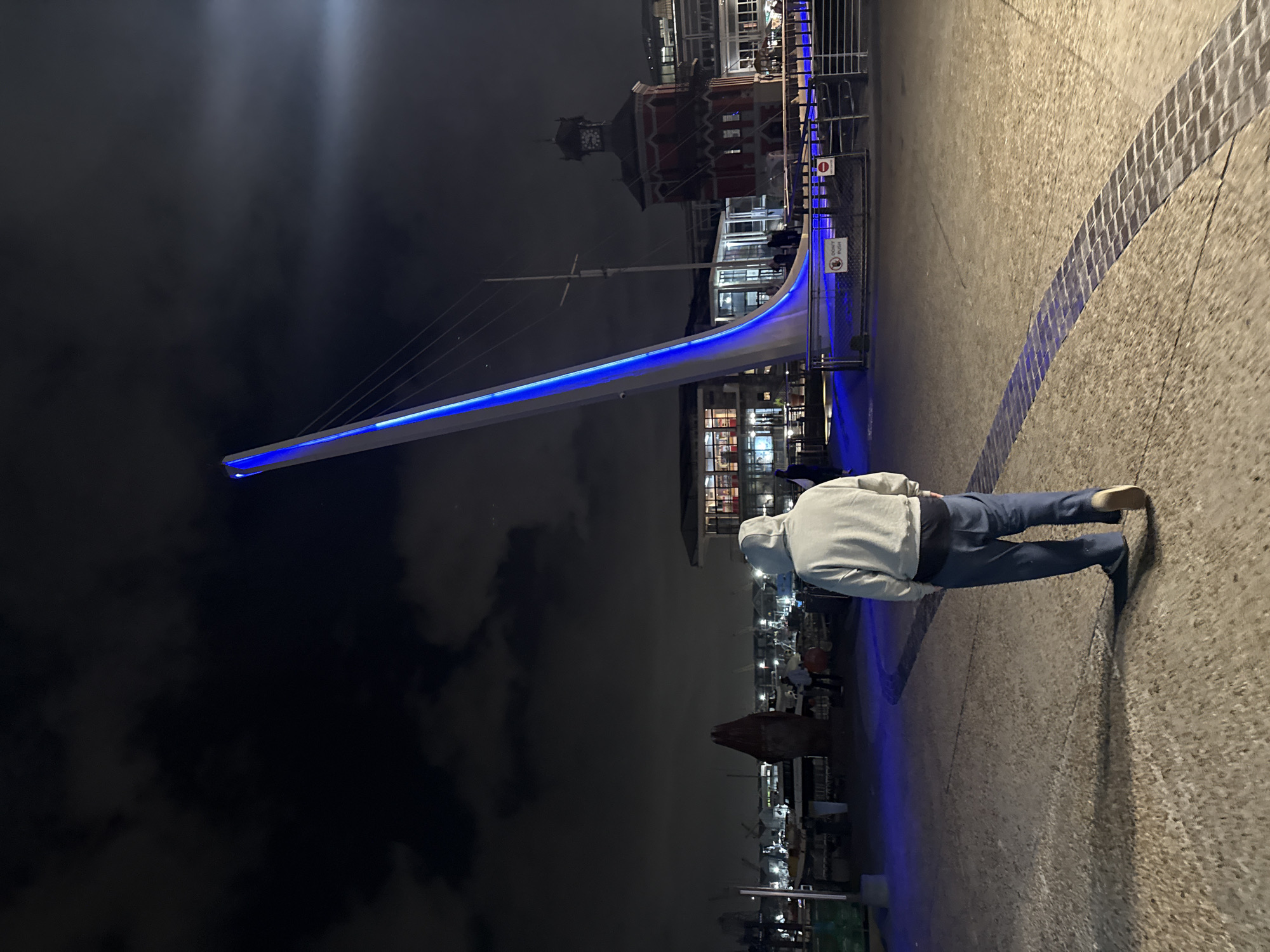 At night in Cape Town, South Africa, a person in a hoodie and jeans walks along a waterfront promenade illuminated with blue light. The bridge, a modern architectural structure, is also illuminated with blue light and stands out against the dark sky. The V&A Waterfront Clock Tower is visible in the background. The scene suggests a calm evening atmosphere.