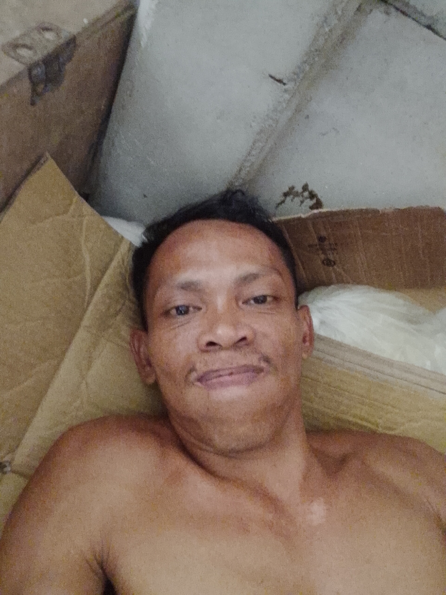A shirtless, brown-skinned man lies in a cardboard box. The photo was taken from above and slightly to the right of his face. The surrounding area includes more cardboard boxes and what appears to be a concrete or stucco-type wall. There is no information within the photo that indicates the exact location of Bukit Ketok, Indonesia.