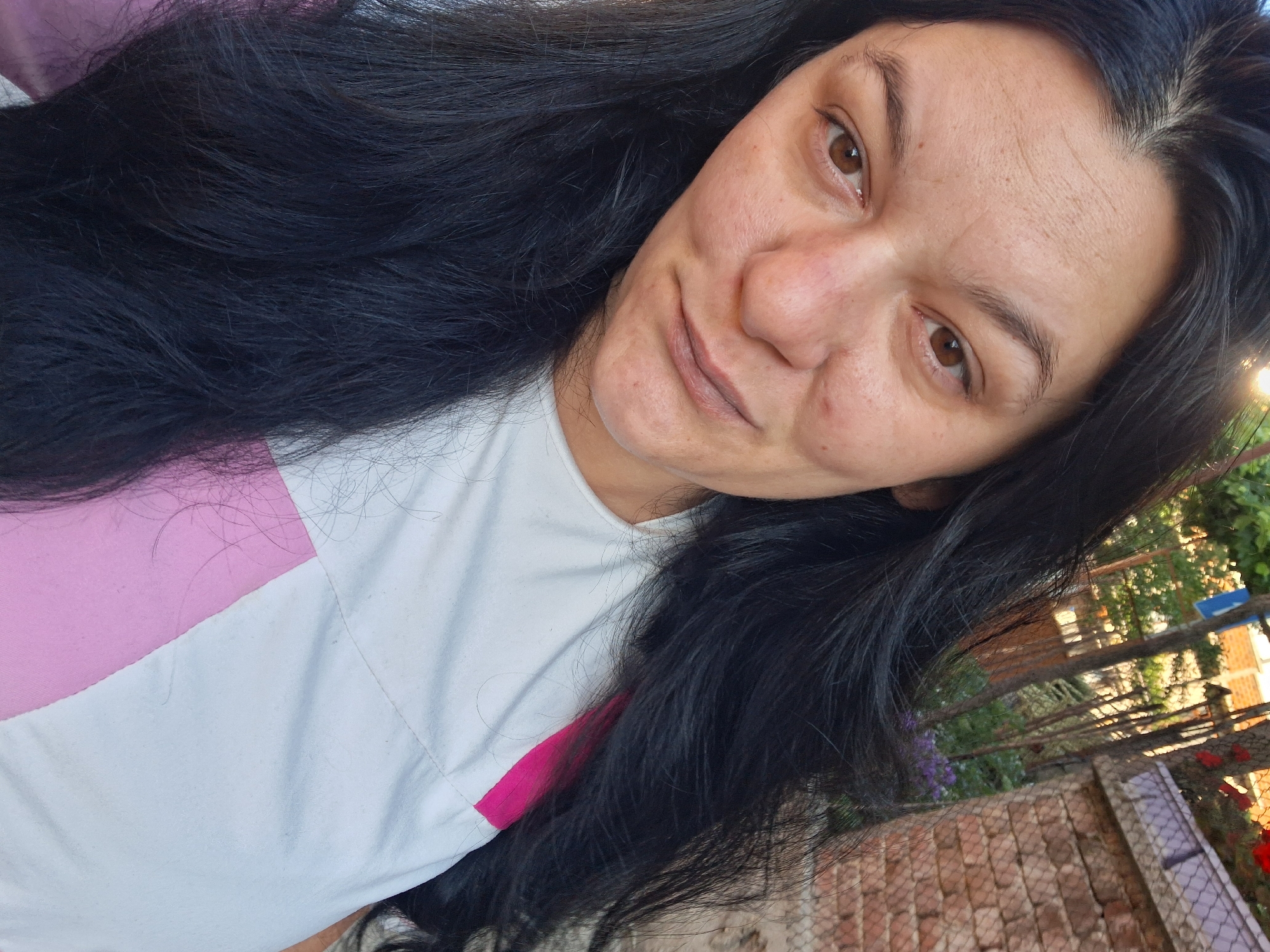 The primary subject is an adult female positioned slightly off-center, gazing directly into the camera. She has long, dark, likely black hair, appearing straight to wavy, which frames her face and extends over her shoulders and chest. Her skin tone is light to medium olive, with visible pores, minor blemishes on her cheeks, and horizontal creases across her forehead. Her eyes are dark brown, and her eyebrows are naturally shaped and dark. Her nose exhibits slight redness at the tip, and her lips are naturally pigmented, forming a subtle, closed-mouth expression. She wears a short-sleeved garment primarily white, featuring a prominent fuchsia or bright pink color-block panel on the right side of her torso (viewer's left). The fabric appears to be cotton or a similar textile, showing some minor textural irregularities.
The scene is set outdoors under direct sunlight, indicated by bright highlights on her hair and the background, suggesting either early morning or late afternoon. The background is partially blurred. To the subject's right (viewer's left), lush green foliage and bright, indistinct light sources are visible. To her left (viewer's right), a weathered red brick wall with visible mortar joints runs horizontally, partially topped by a chain-link fence. Sparse green vegetation, including tree trunks and small clusters of purple flowers, is present behind the fence. Distant, out-of-focus residential structures with reddish roofs and a faint blue sign are discernible in the upper background. The overall environment suggests a residential area or garden in Gotse Delchev, Bulgaria.
