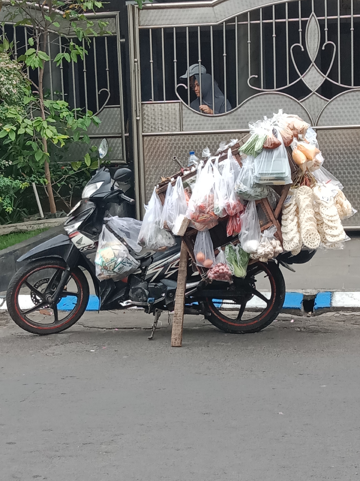 The image depicts a motorcycle laden with various goods, suggesting it's used for mobile vending. The motorcycle is parked on an asphalt road, with a blue and white striped curb to its right. Behind the motorcycle is a metal gate and a building facade. A person, partially obscured by the gate, appears to be looking out from within the building. The motorcycle is carrying an assortment of items in plastic bags, including what appear to be vegetables, onions, and crackers, along with a scale. The setting is identifiable as being in the city of Ngingas, Indonesia, based on the provided context. The overall scene conveys a sense of everyday life and commerce in an Indonesian urban environment.