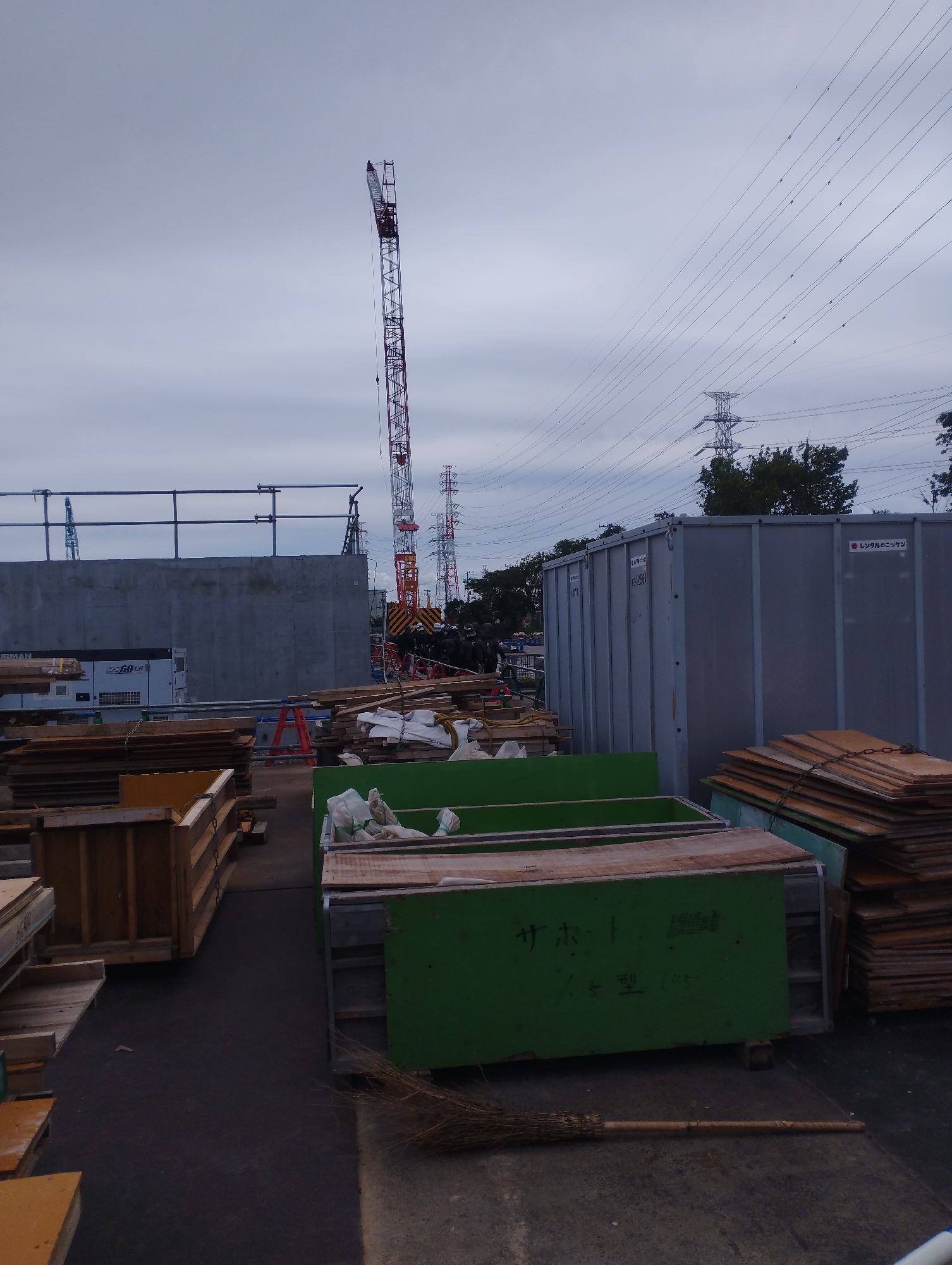 A construction site in Ichikawa, Japan features a tall red and white tower crane, a grey shipping container, stacks of wood, and various construction materials. In the background, electrical towers and power lines crisscross the overcast sky. The scene is devoid of people and shows no active construction work. The image captures a still moment in the ongoing development of the area.