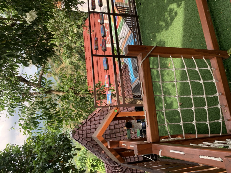 The image shows a wooden playground structure with a rope net. The structure is located on a rooftop with a view of the surrounding forest. There is a small child standing near the edge of the platform wearing a white shirt and blue shorts. The child is looking towards the left of the image. In the background, there is a red roofed building with a metal railing that is covered with various colorful packages. Behind the building, there is a swimming pool that is mostly out of view. There is a green artificial grass surface on the rooftop. The image was taken in Kecamatan Borobudur, Indonesia (latitude: -7.606, longitude: 110.18).