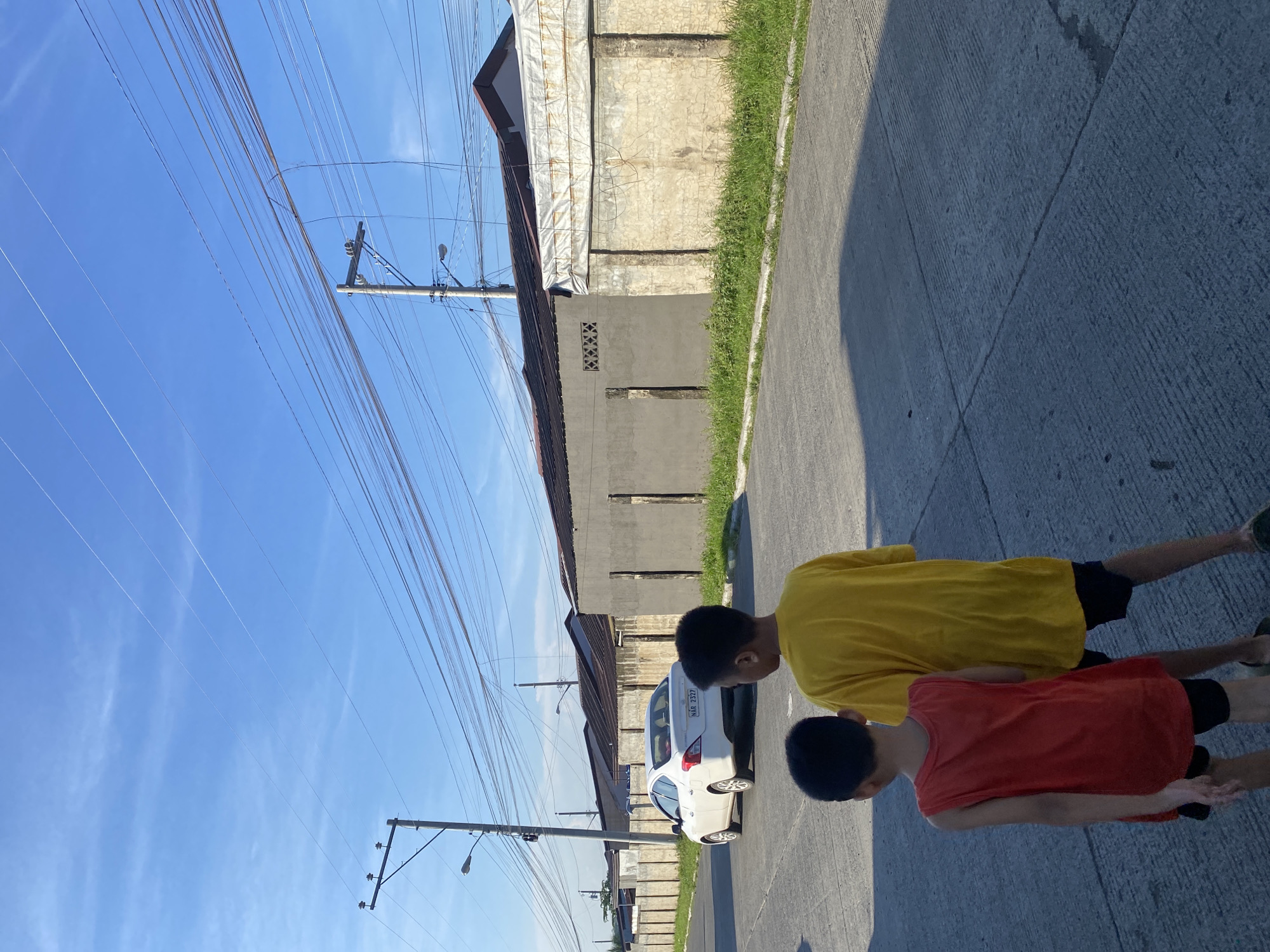 The image depicts a street scene in Davao City, Philippines, with two young boys walking away from the camera. The boy in the foreground wears a red sleeveless shirt and black shorts, while the boy behind him is dressed in a yellow t-shirt and black shorts. They are walking on a concrete road, casting long shadows from the overhead sun. A white sedan is parked on the left side of the road, facing away from the viewer. In the background, a line of concrete walls borders the road, topped with corrugated metal roofing. Overhead, a complex network of utility wires crisscrosses against a bright blue sky with scattered thin clouds. The overall impression is a typical residential street in a warm, sunny climate.