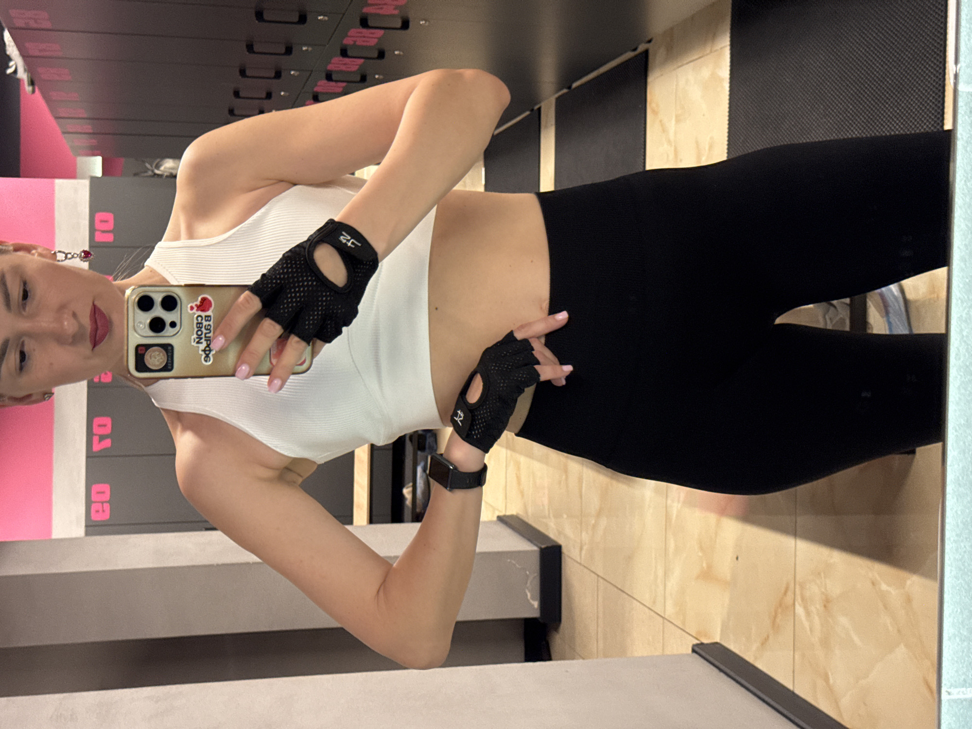 A woman with blonde hair, wearing workout gloves, a white tank top, and black leggings, takes a selfie in what appears to be a gym locker room. She holds a gold-colored iPhone, which has a sticker on it, up to her face. Lockers are in the background, some with pink accents, and there is also some light-colored tile visible. The location is potentially in Sochi, Russia, as the sticker on the phone is written in Russian.