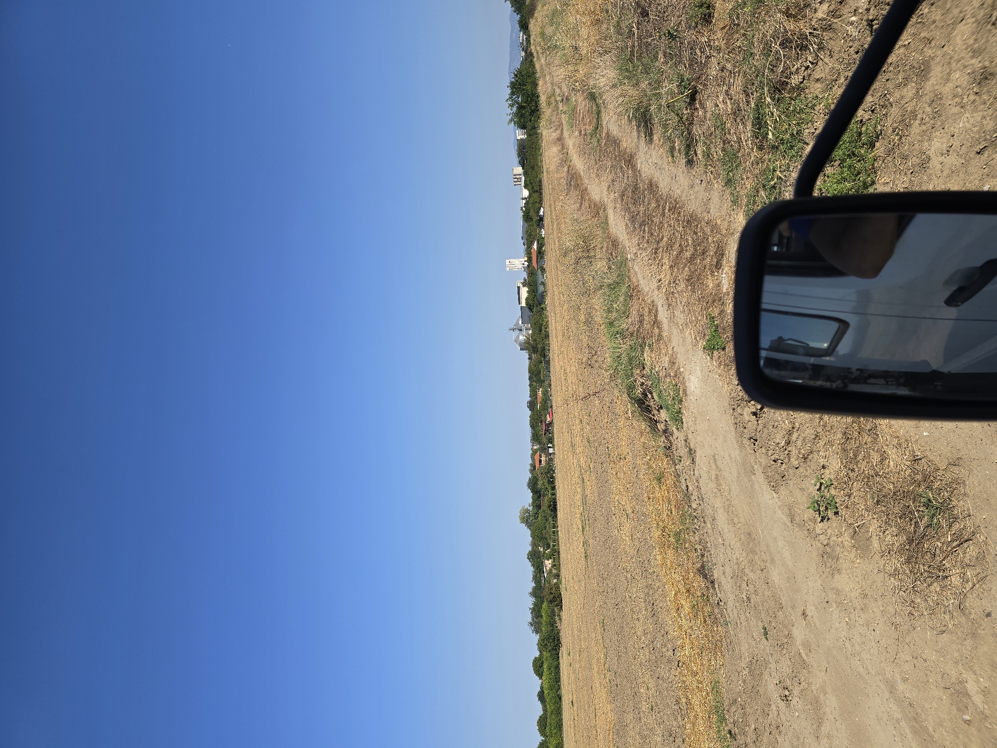 An outdoor, daytime, eye-level, vertical shot captures a dirt road traversing a dry, harvested field under a clear blue sky. The foreground features the side-view mirror of a white vehicle, reflecting the sky and the interior of the vehicle, including a person's arm and shoulder. The road, marked by tire tracks, winds through the parched earth and dried grass. In the mid-ground, a line of trees separates the field from a cluster of buildings in the distance. These buildings appear to be industrial or agricultural, with silos and tall structures visible. The scene is likely situated in a rural or semi-rural area surrounding Yambol, Bulgaria, given the presence of agricultural land and industrial facilities characteristic of such regions. The overall impression is one of a dry, sunny day in an agricultural landscape.