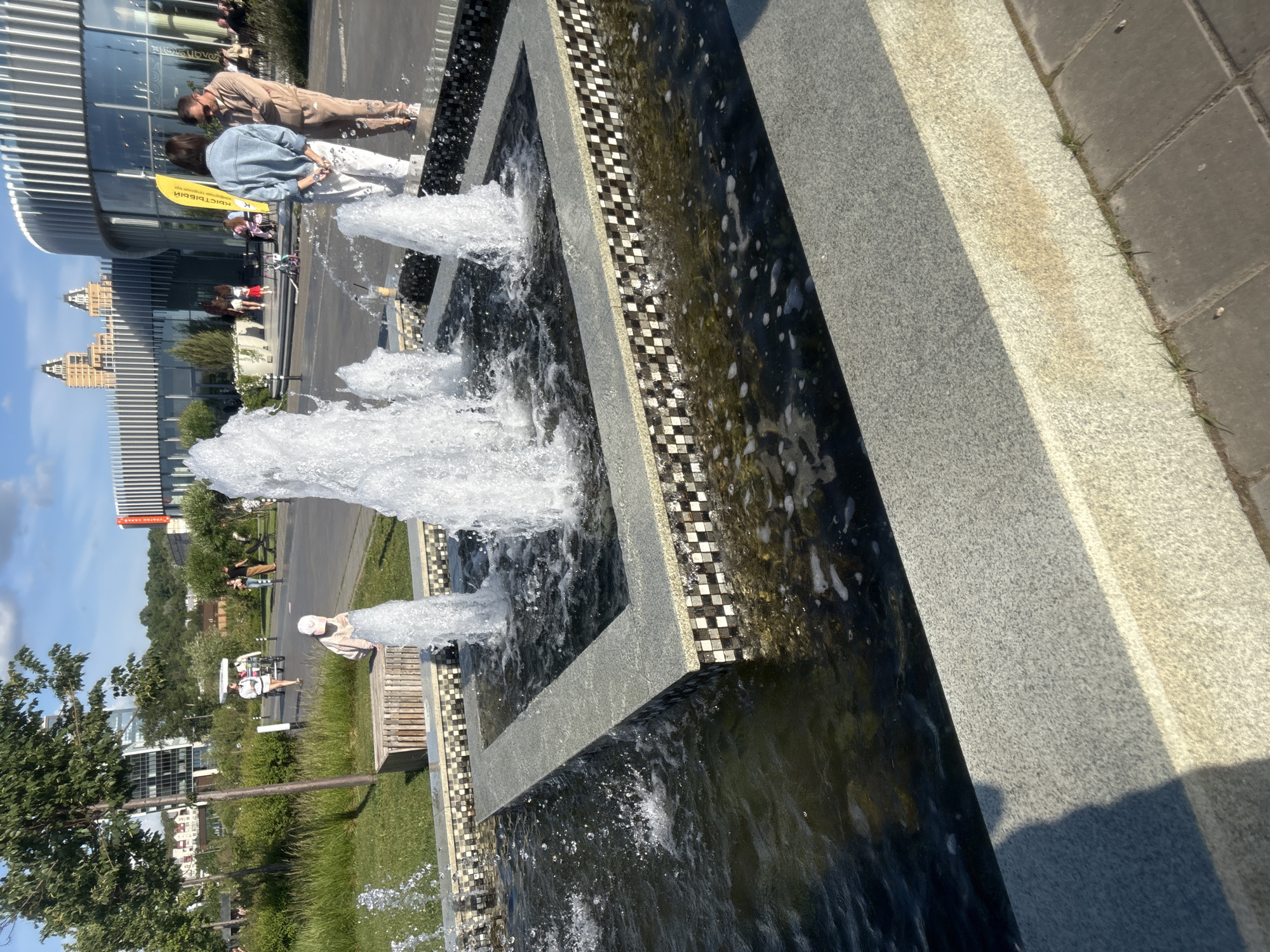 This image captures a vibrant public space in Kazan, Russia, under bright, clear daylight, likely in the late morning or afternoon due to the angle of shadows.
In the foreground, a multi-tiered fountain is prominently displayed. It features rectangular granite borders in light grey, with its vertical faces adorned by a checkered mosaic pattern of black, white, and tan tiles. Water jets vigorously upwards from multiple nozzles within the dark, reflective basin, creating white foam and splashes. The immediate ground in front of the fountain consists of light grey speckled paving, exhibiting a strong shadow from an unseen source on the left; this transitions to darker grey rectangular pavers with minimal grass growth on the right.
The midground and background reveal an active urban environment. To the left, a paved path meanders through a landscaped area featuring green grass, trees, and shrubs. A young child, possibly female, in a light-colored cap and top, stands near a wooden bench beside this path, facing the fountain. Further along the path, a woman in a white top and shorts pushes a white three-wheeled stroller or cart. In the background, modern buildings with glass facades and residential structures are discernible through the greenery.
To the right, a distinctive circular building with a predominantly glass facade and vertical white architectural accents serves as a backdrop. An outdoor cafe area with tables and chairs is positioned in front of it. Two adults stand near the fountain: a woman, viewed from behind, wearing a light blue denim jacket and white patterned pants with her hands clasped behind her back; and a man, facing her, dressed in a beige tracksuit and sunglasses, looking downwards. A yellow vertical banner with partially visible Cyrillic text reading "КЫИСТЫЙ К..." stands between them and the building. In the far background, two prominent multi-story residential towers with dark, angular upper sections are visible against a bright blue sky with sparse clouds.
