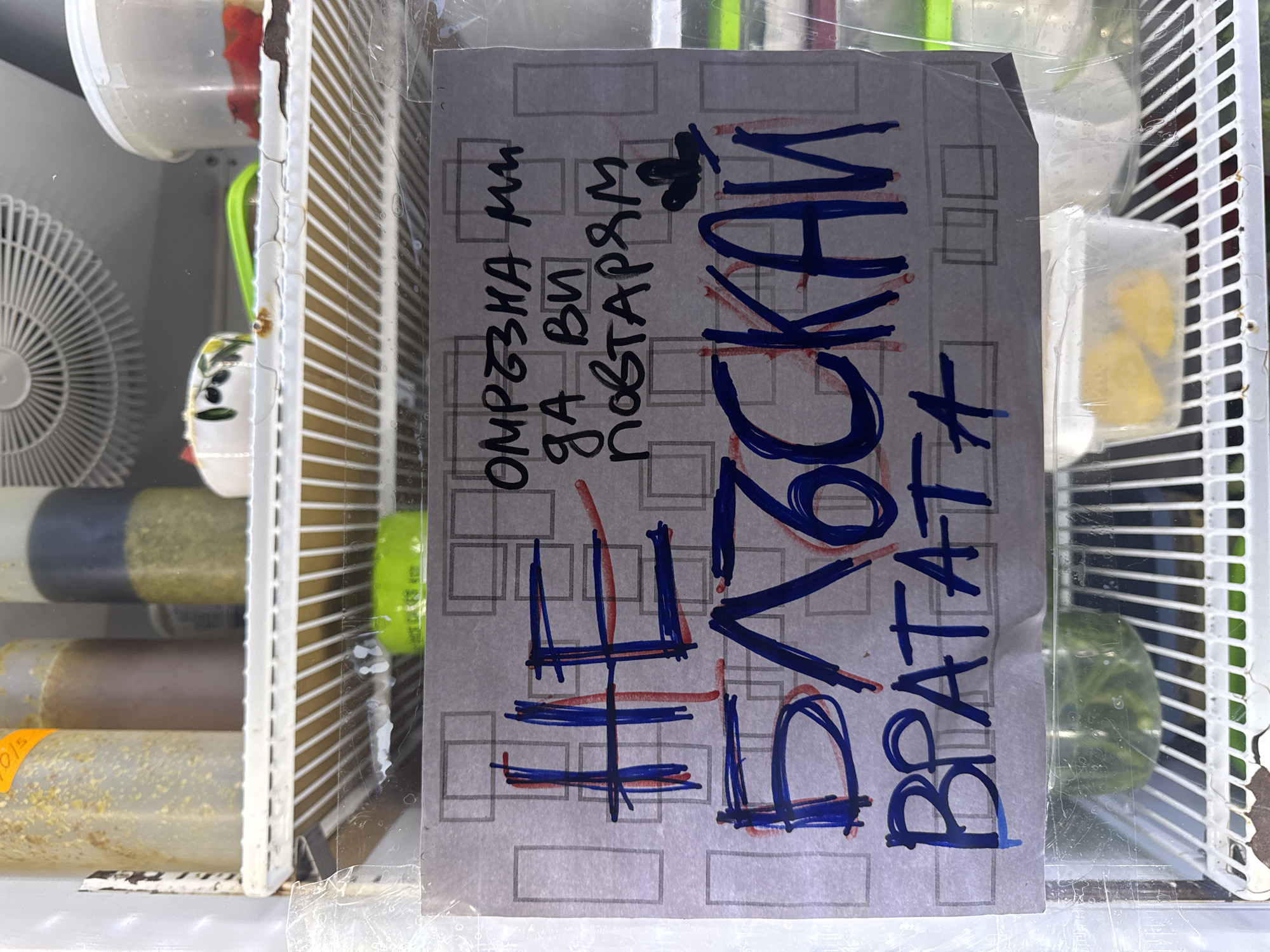 This media file captures an eye-level view inside a refrigerator or cooler, likely in a shop or cafe in Sofia, Bulgaria. The main subject is a handwritten sign, taped to an inner surface, covered in protective plastic film.
The sign features Cyrillic text, with some words in black marker and others aggressively emphasized and partially crossed out in blue and red. The black text reads "ОМРЪЗНА ми ДА ВИ ПОВТАРЯМ", which translates to "I'm tired of telling you". Below this, large, bold letters, heavily outlined and crossed out in blue and red, spell out "НЕ ПИПКАЙ", meaning "DO NOT TOUCH". Beneath this, in large blue letters, is "ВРАТАТА", which means "THE DOOR". The overall message conveyed is a frustrated plea: "I'm tired of telling you, DO NOT TOUCH THE DOOR".
Behind and around the sign, the interior of the refrigerator is visible. On the left, a white fan unit can be seen. To the right and below, various clear plastic containers and bottles are arranged on white wire shelves. These containers hold different liquids and food items, including bottles with green, brown, and light yellow liquids (possibly juices, smoothies, or sauces), and small clear boxes containing yellow fruit chunks. The contents suggest that the refrigerator stores food or beverages for sale. The lighting is bright, indicating an indoor setting. The tone of the sign suggests a recurring issue with customers touching the refrigerator door.
