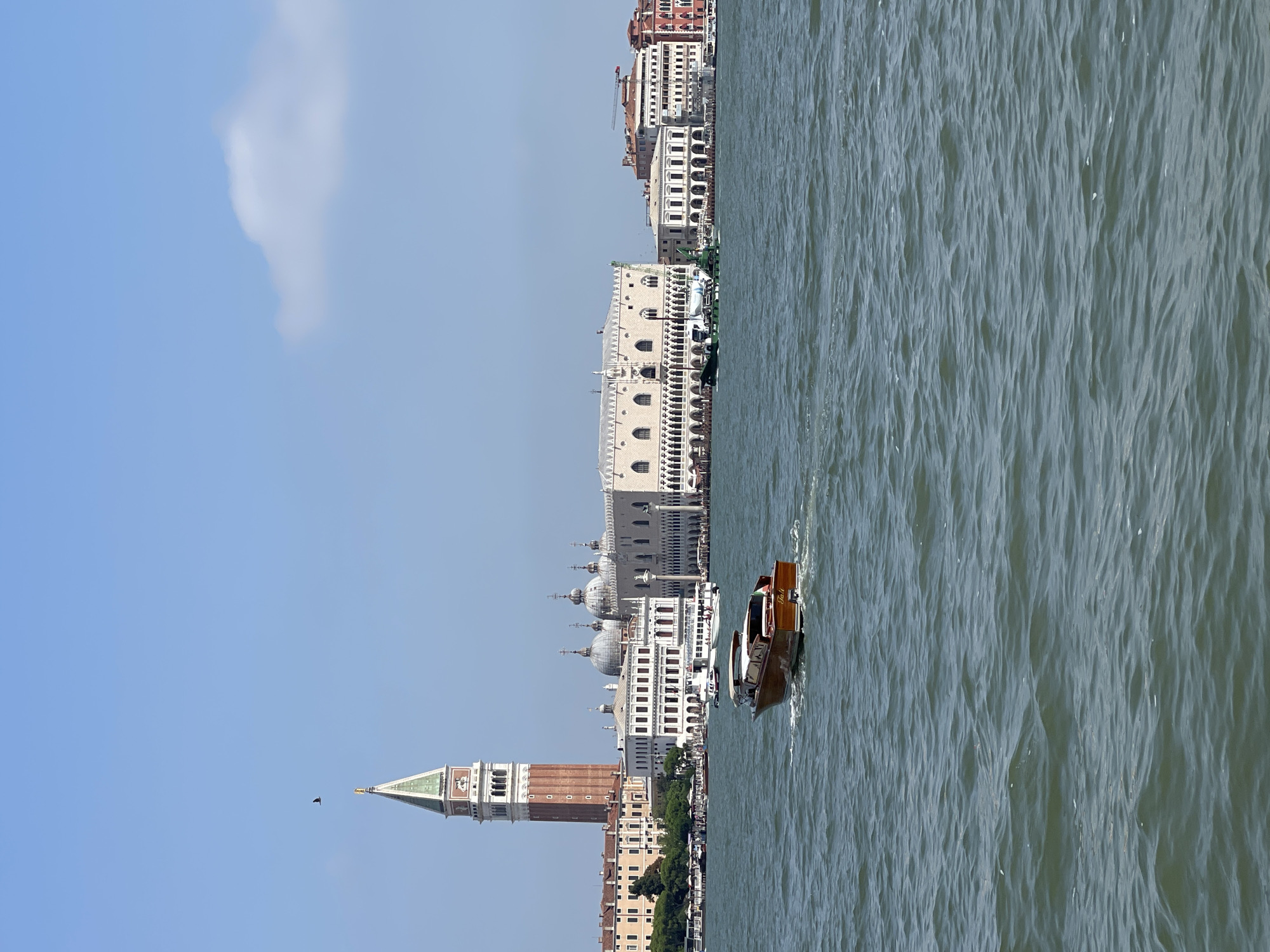In a wide shot overlooking a body of water, a brown boat with a white canopy cuts through the choppy blue-green surface, leaving a white wake. In the background, the city skyline features a tall, red brick bell tower with a green spire and clock face. Behind the bell tower, a long, ornate white building with arched windows and a red-tiled roof stretches across the scene. Other buildings, some with similar red roofs, line the waterfront. The sky is a clear blue with a few wispy white clouds.