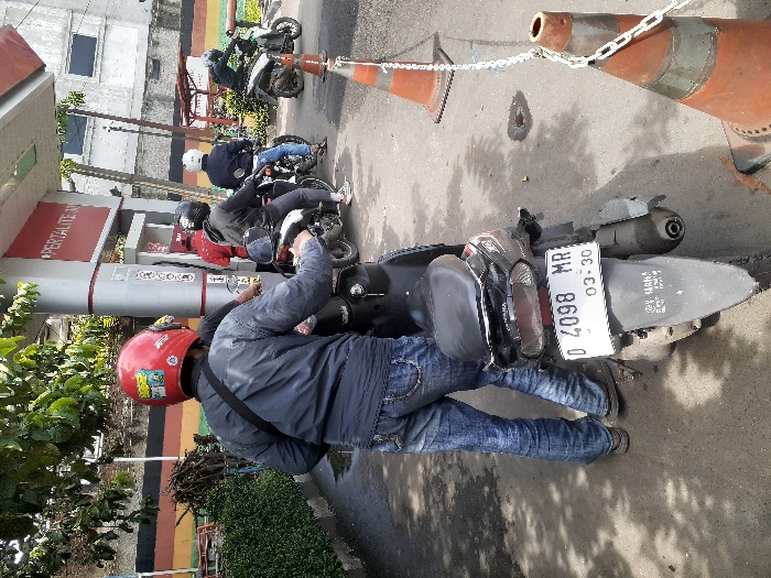 The image shows a man wearing a red helmet standing next to a black scooter with the license plate "D 4098 MR 03-30". The scooter is parked on a paved road in front of a gas station at the location of latitude: -6.863, longitude: 107.51 in Ngamprah, Indonesia. Behind the scooter are two traffic cones. In the background, there are two men refueling their motorcycles at the gas station. The man in the red helmet is holding onto the handlebars of the black scooter, while the other man is on his phone. There are trees in the background.