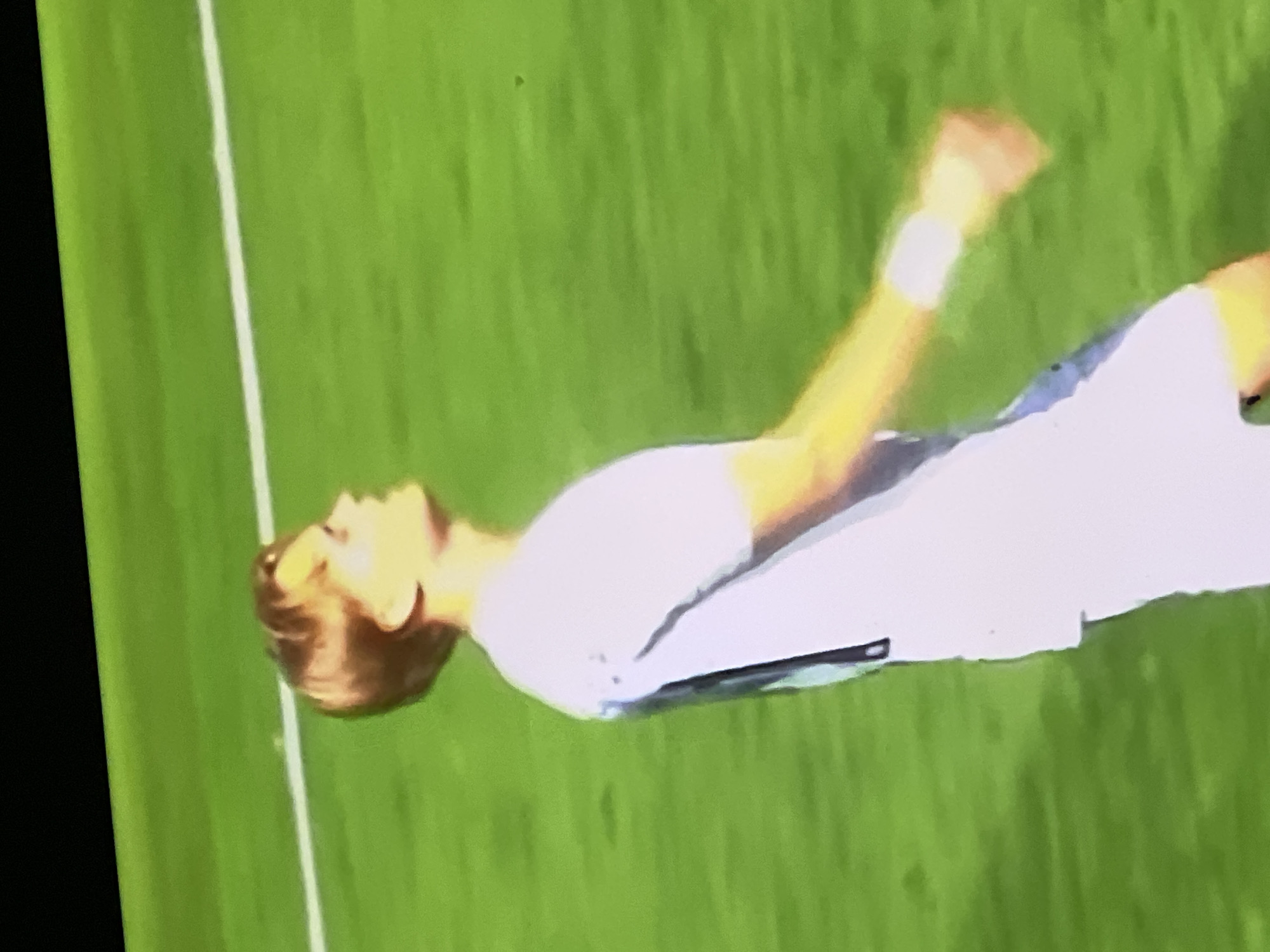 The image depicts a young male, seen from the chest up in profile, standing on a verdant green sports pitch. He possesses light brown hair and fair skin, dressed in a plain white short-sleeved shirt and a white wristband on his right arm. His gaze is directed upwards and to his right, and his right arm is bent at the elbow. A distinct white line, characteristic of a playing field, is visible across the green surface behind him. A dark, undefined structure occupies the top left of the frame. This scene is located in the city of Casablanca, Morocco.