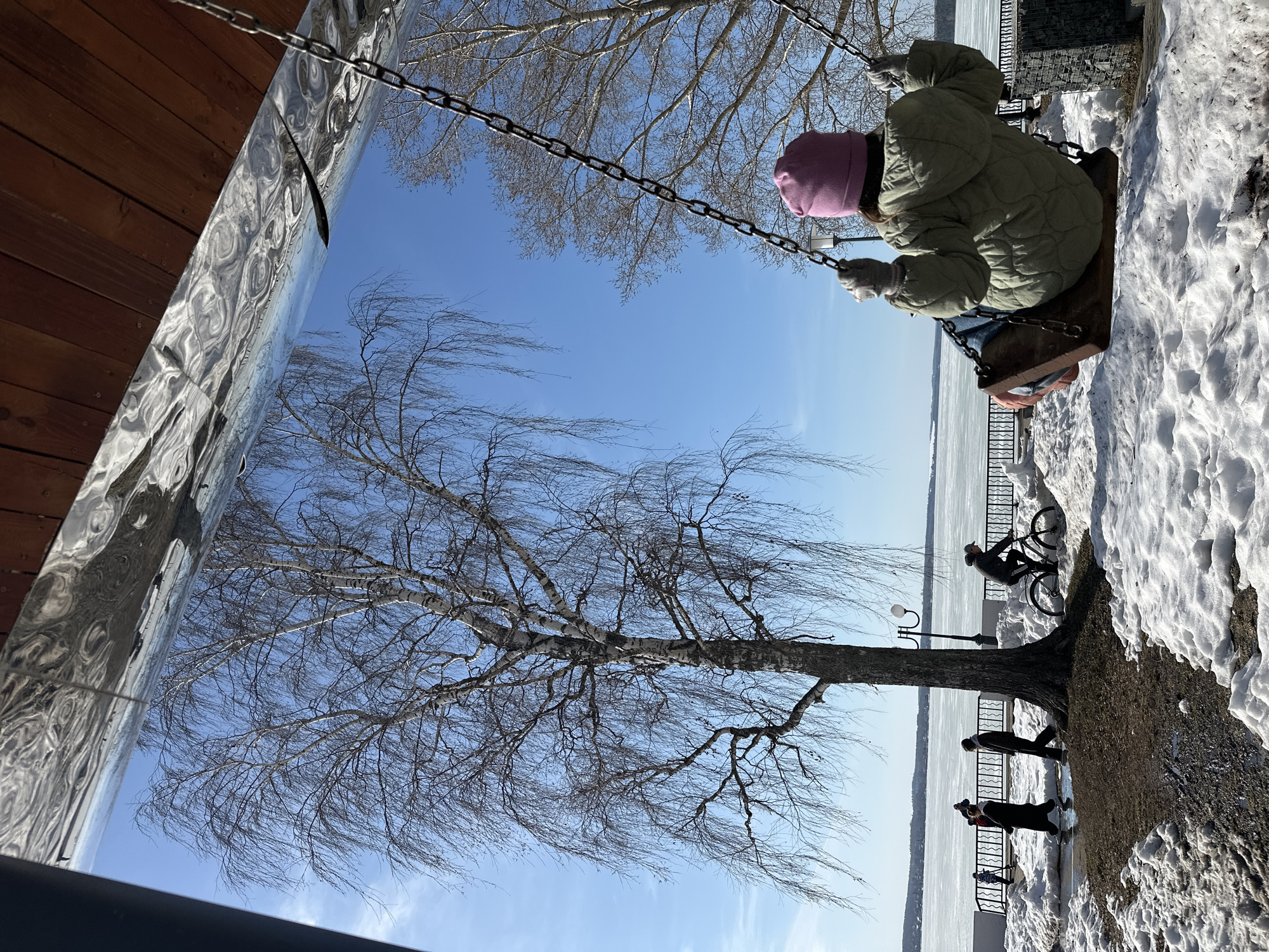 A person wearing a pink hat and green jacket sits on a swing. The swing hangs from chains connected to a structure. The location appears to be a waterfront area in the city of Votkinsk, Russia. In the background, a frozen body of water (likely a lake) stretches to the horizon. The shoreline is visible with a railing. Some people are walking along the waterfront path, while another person is riding a bicycle along the path. A tree with bare branches stands near the path. Snow is on the ground on the side nearest the swing. Part of the structure the swing is attached to reflects the tree and sky. The sky is blue with a few clouds.