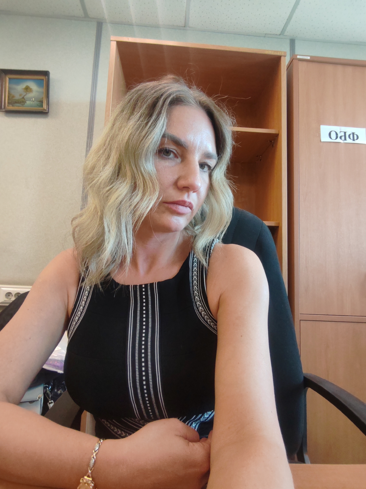 A woman is sitting at a desk, indoors. She is wearing a black tank top with white accents. The woman is fair-skinned with light-blonde, wavy hair. A piece of jewelry is on her wrist. The background includes office furniture, specifically a wooden cabinet and a wooden shelf, along with an indistinct picture hanging on the wall. The environment is a typical office setting. There is no evidence to conclude this image was taken in Petropavlovsk-Kamchatsky, Russia.