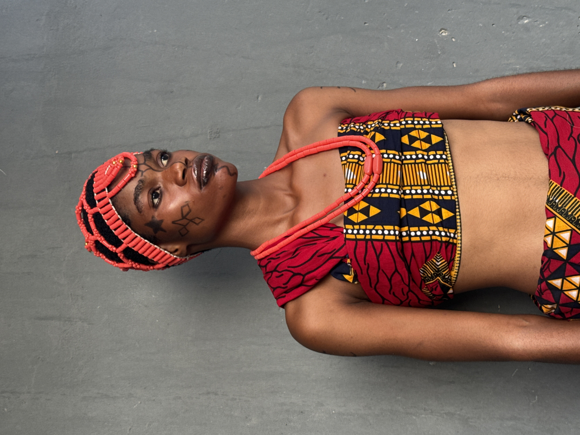 The image shows a young woman lying on a gray surface. She is wearing a traditional African outfit with a red and black patterned top and bottom, and a red beaded headdress and necklace. She has tribal markings on her face. The background is a plain gray color. It is not possible to determine the exact location in Badagry, Nigeria from the image alone.