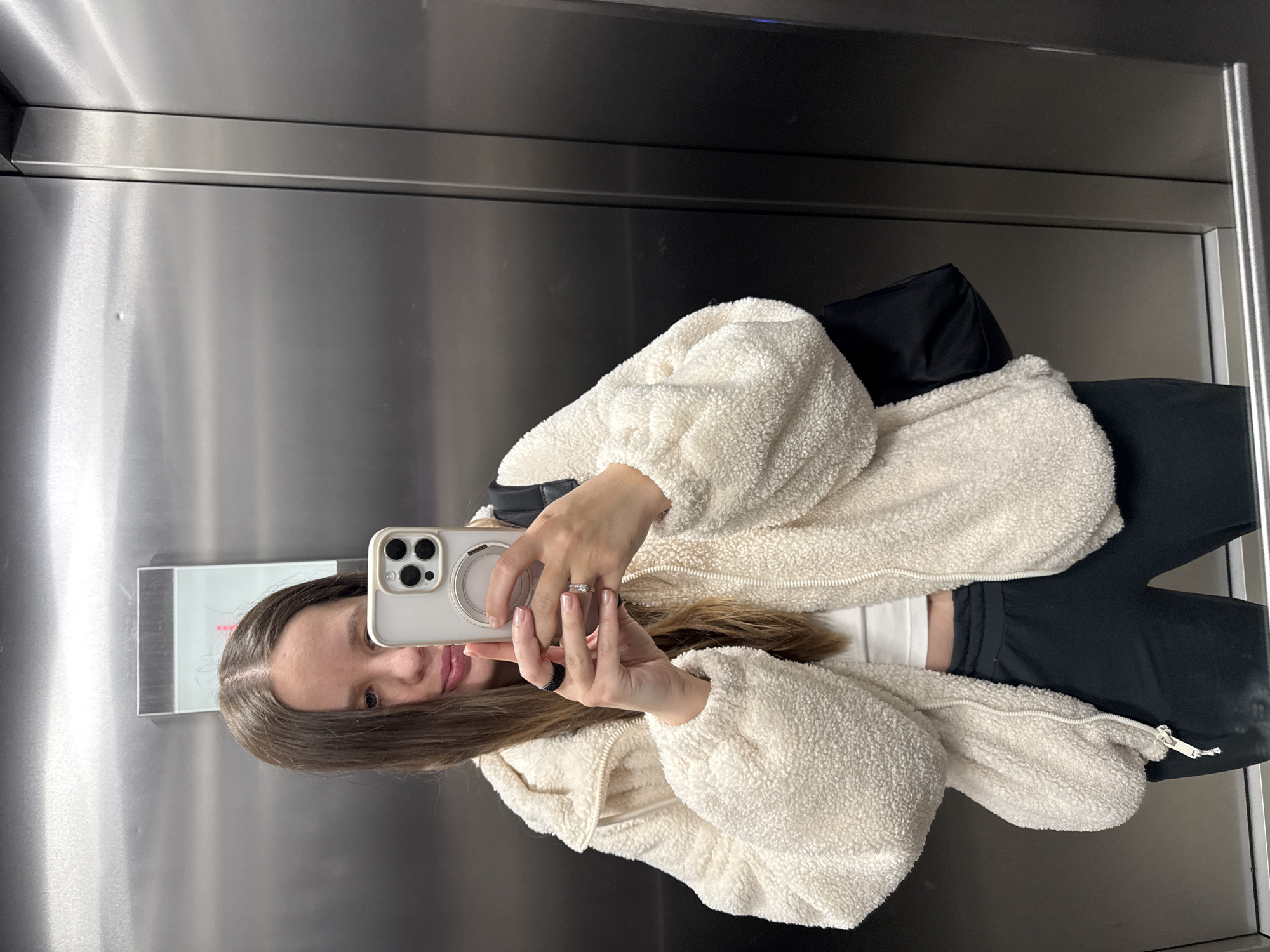 A person is taking a selfie in what appears to be the interior of an elevator. The elevator walls are metallic, and a digital display panel is partially visible to the left of the person's face, reflected in the mirror. The individual is holding a smartphone to take the photo, wearing a white, fluffy jacket and dark-colored bottoms. A dark-colored bag is also visible behind the jacket. There are no indicators or clues to establish a definitive location in Barcelona, Spain.