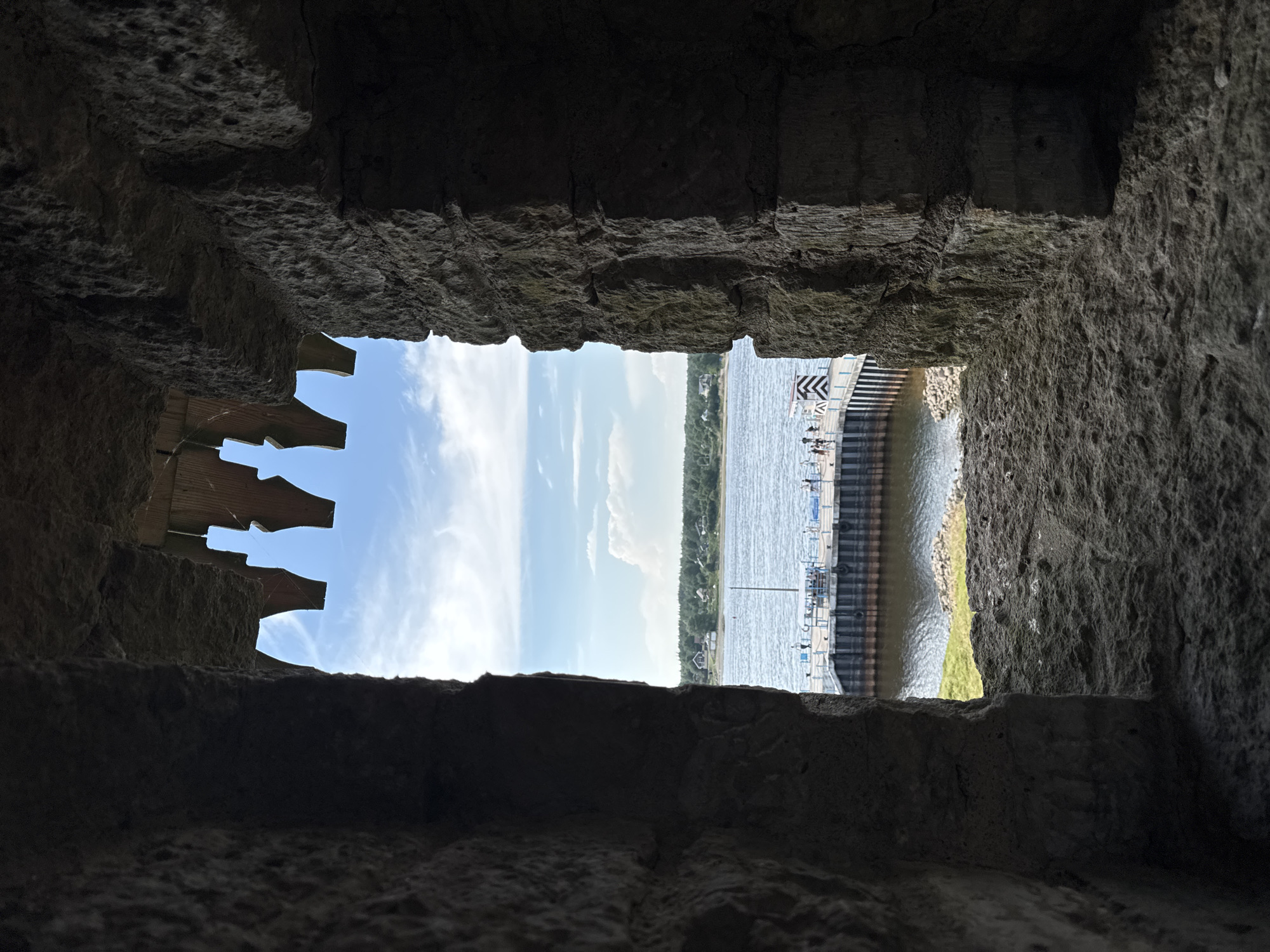 This image shows a view through a rectangular opening in a stone structure, possibly a castle or ruin, looking out onto a body of water under a bright, cloudy sky. The opening is framed by rough, dark stone walls, creating a strong contrast with the scene beyond.
Through the opening, a pier or dock extends into the water. Several figures, appearing as small silhouettes, are visible on the pier. The water is calm and reflects the sky. On the far shore, a line of trees borders the water, with a few buildings interspersed among them. The sky is a bright blue with scattered white clouds, suggesting it is daytime, likely mid-morning or afternoon given the lighting.
On the left side of the opening, a wooden structure with a jagged, decorative edge is visible, partially obscuring the view of the sky and water. This detail adds an element of mystery to the scene. The overall impression is one of looking out from a protected, ancient space onto a vibrant, active waterfront. The location context of Shlisselburg, Russia, suggests this might be a view from the Oreshek Fortress, a historic site on an island in Lake Ladoga, known for its stone architecture.