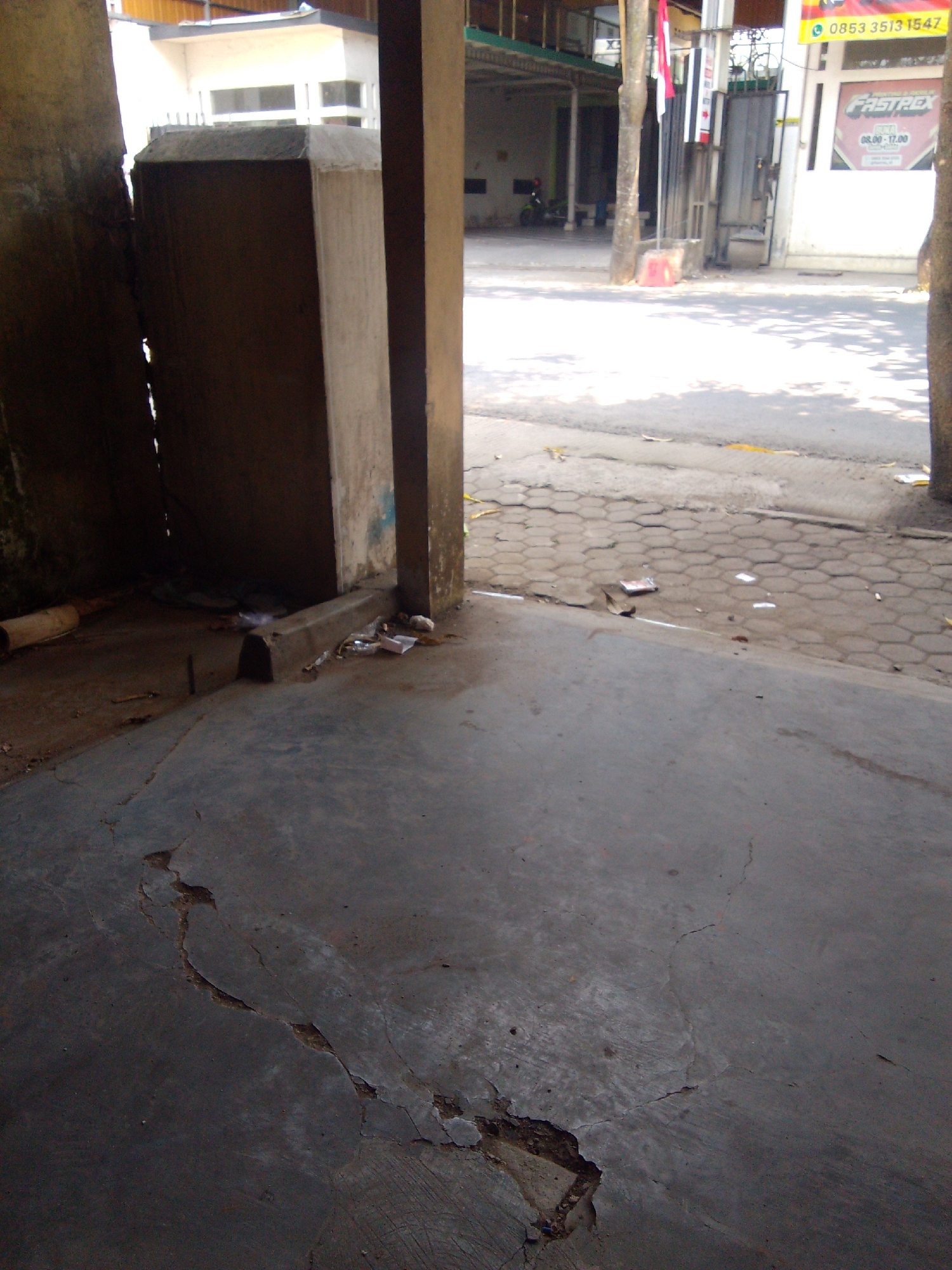 The image depicts an outdoor scene, likely in an urban or semi-urban environment. In the foreground, a cracked concrete surface dominates, showing signs of wear and tear. A low concrete barrier runs along the edge of this surface. To the left, a rough, textured vertical surface, possibly a wall or a pillar, is visible. Further back, through an opening, a paved area with a hexagonal pattern is seen, with scattered debris, including paper and possibly plastic. Beyond this, a road or street is visible, with sunlight casting shadows. On the right side of the image, a modern building with large windows and a prominent sign with the text "FASTREX" and "08.00-17.00" is partially visible. A red and white Indonesian flag is also displayed near this building. A motorcycle is parked under a covered area in the background. The overall impression is one of a somewhat neglected public space adjacent to commercial establishments. The presence of the Indonesian flag and the building signage suggests a location within Indonesia. Given the reference to Trenggalek, Indonesia, this scene is situated in that city.