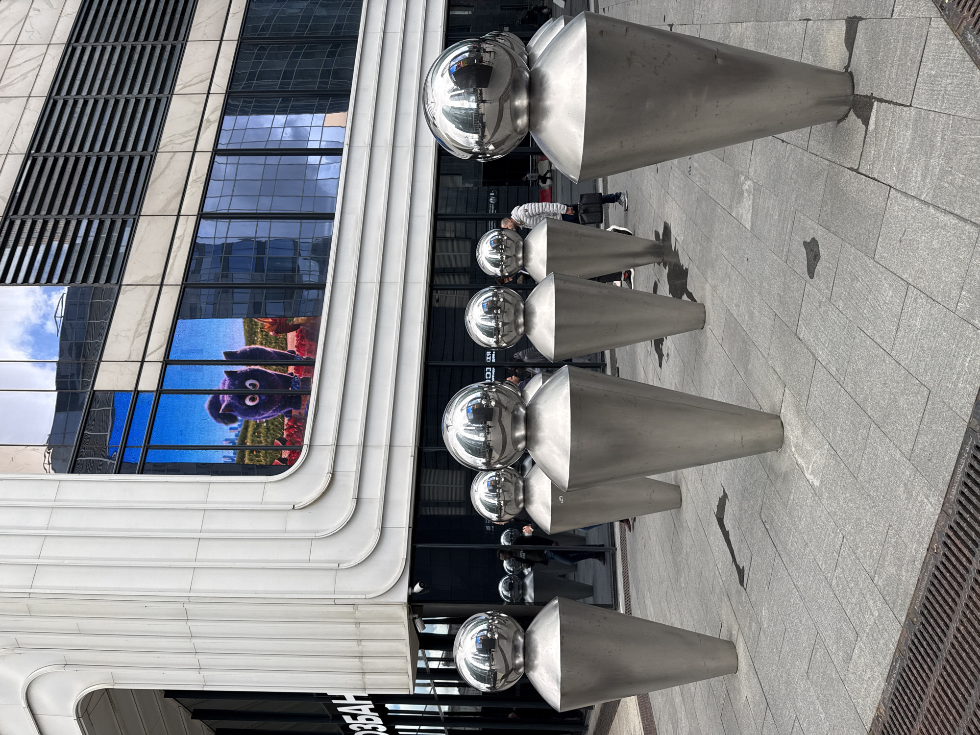 This is a photograph of the business district of Moscow, Russia, specifically depicting a public art installation in front of a modern glass-facaded building. The art installation consists of several large, silver, conical sculptures topped with spherical elements, arranged in a row on a paved plaza. Some of the sculptures appear to be reflecting the surrounding environment, including the sky and other buildings. In the background, the building features a large window displaying a digital image of a cartoon-like purple cat with big eyes looking out at the viewer. People are visible walking in the background, some interacting with the plaza and sculptures, while others are walking past. The plaza has a grid-like pavement pattern and a metal grate along the edge. The lighting suggests it is daytime, with some overcast conditions as evidenced by reflections of clouds in the building's windows.