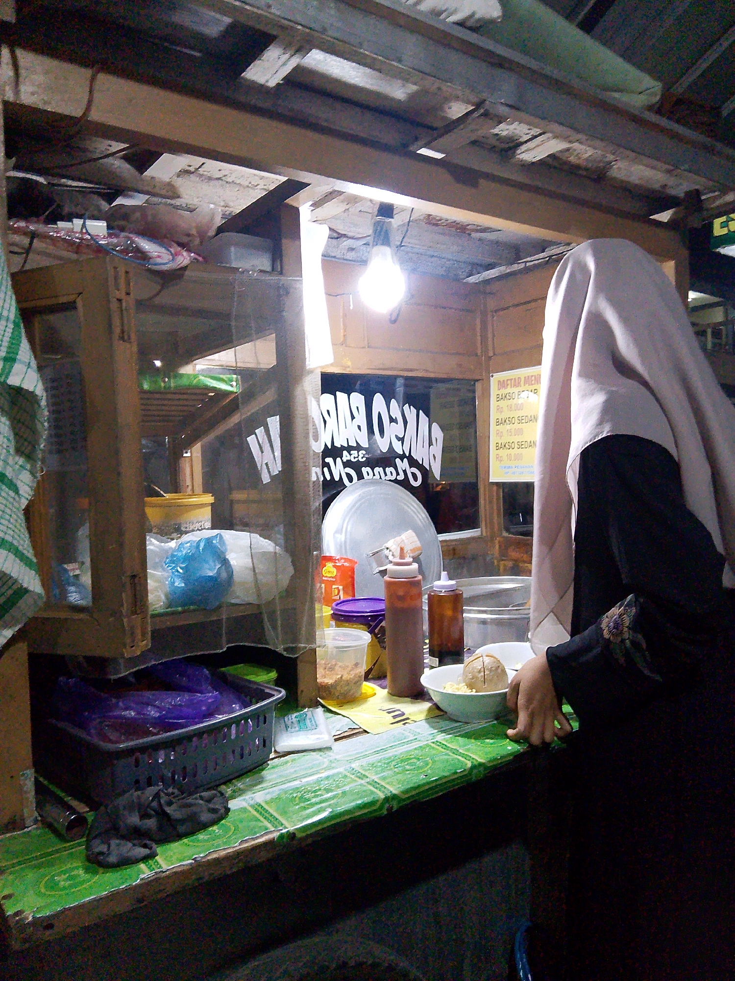 The image depicts a street food stall, possibly located in Wanareja, Indonesia, judging by the "BAKSO SEDAN" sign. A person wearing a light pink hijab and a black garment is partially visible, appearing to be serving or preparing food. The stall features a wooden counter with a green, tiled surface. On the counter, there are several items including a bowl of noodles with a large meatball, two condiment bottles, a plastic container with food, and other unidentified objects. Behind the counter, shelves hold various food items and containers, including yellow buckets and blue plastic bags. A light bulb hangs overhead, illuminating the scene. A sign board displays menu items and prices in Indonesian currency. The overall setting suggests a bustling, informal food business operating in an outdoor or semi-outdoor environment.