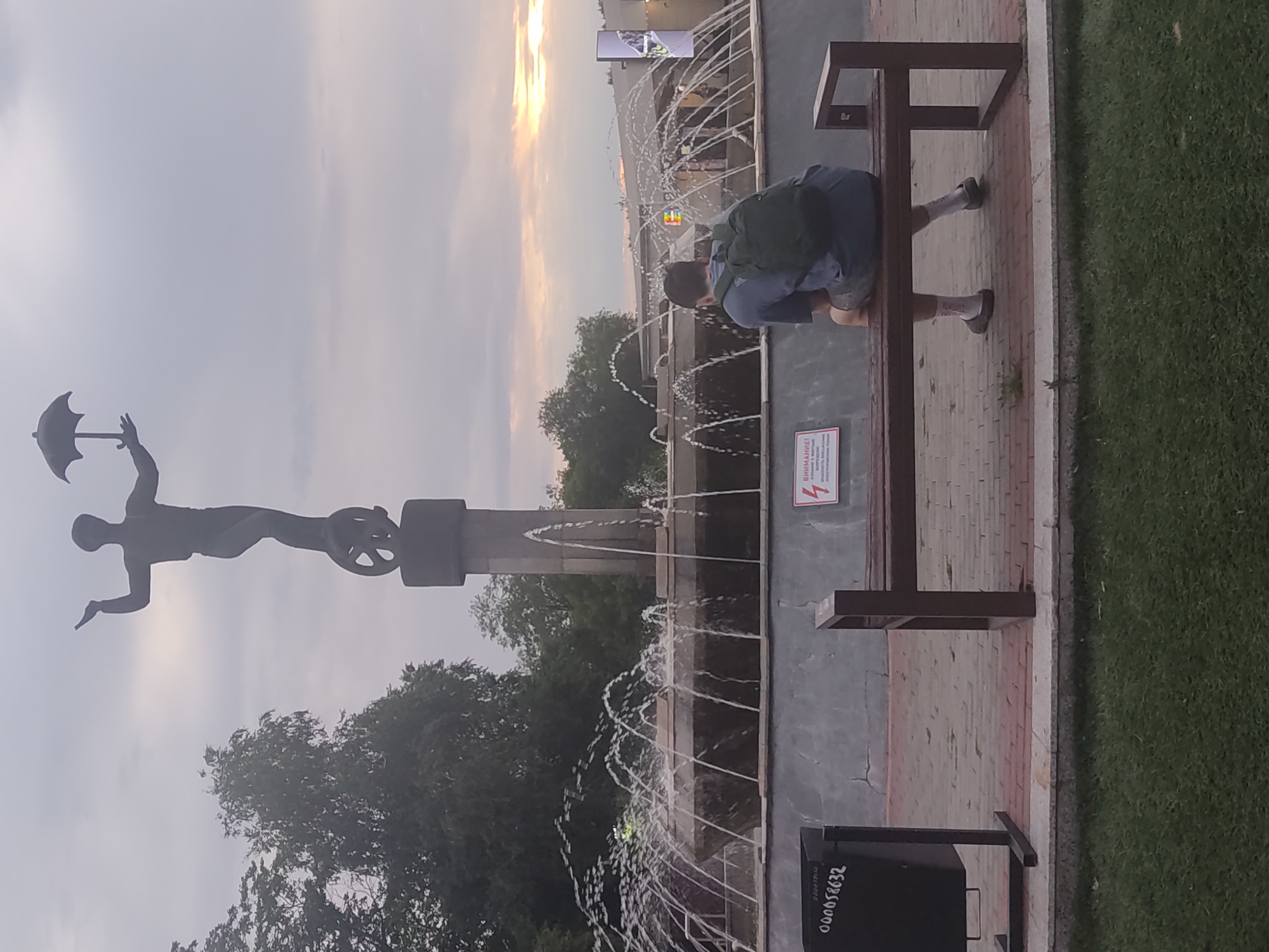 A person is crouching on a park bench next to a fountain. The person is wearing a green backpack and dark clothing. The fountain has several jets of water spraying upwards and outwards. In the background, there is a statue of a person holding an umbrella. The sky is overcast with some hints of sunset colors. The scene appears to be in a public park or plaza. There is a sign on the wall behind the bench with a lightning bolt symbol and some text. The text on the sign appears to be in Cyrillic. There is also a trash can to the right of the bench with some numbers printed on it. The image is oriented vertically.