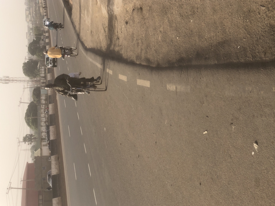 The image depicts a daytime street scene on an asphalt road in Hanwa, Nigeria, under a hazy, light-grey sky. The road surface is light brown-grey with scattered debris. White dashed lines delineate the right edge of the paved road, which transitions to an unpaved dirt shoulder on the right.
In the foreground, a male individual dressed in dark clothing (possibly a dark agbada or similar garment) rides a dark-colored bicycle. He carries a white object, possibly a bag, on the front of his bicycle, and a dark bag or garment over his left shoulder. He faces forward, moving away from the viewer at a slight angle. Long shadows are cast from the right side of the frame onto the road surface.
Further down the road, a second male individual, wearing a bright yellow long-sleeved shirt and dark trousers, rides a bicycle away from the viewer. A white item is secured to the rear rack of his bicycle. Behind him, a third individual rides a motorcycle, also moving away. In the distant background, several light-colored cars are visible on the multi-lane road.
The background features a concrete barrier separating the road from various structures. To the left, a large, reddish-brown building with a flat roof is visible. Beyond the barrier, several smaller buildings with white walls and dark roofs are present, along with green trees and utility poles. A tall, grey metal telecommunications tower stands prominently against the hazy sky in the far background.