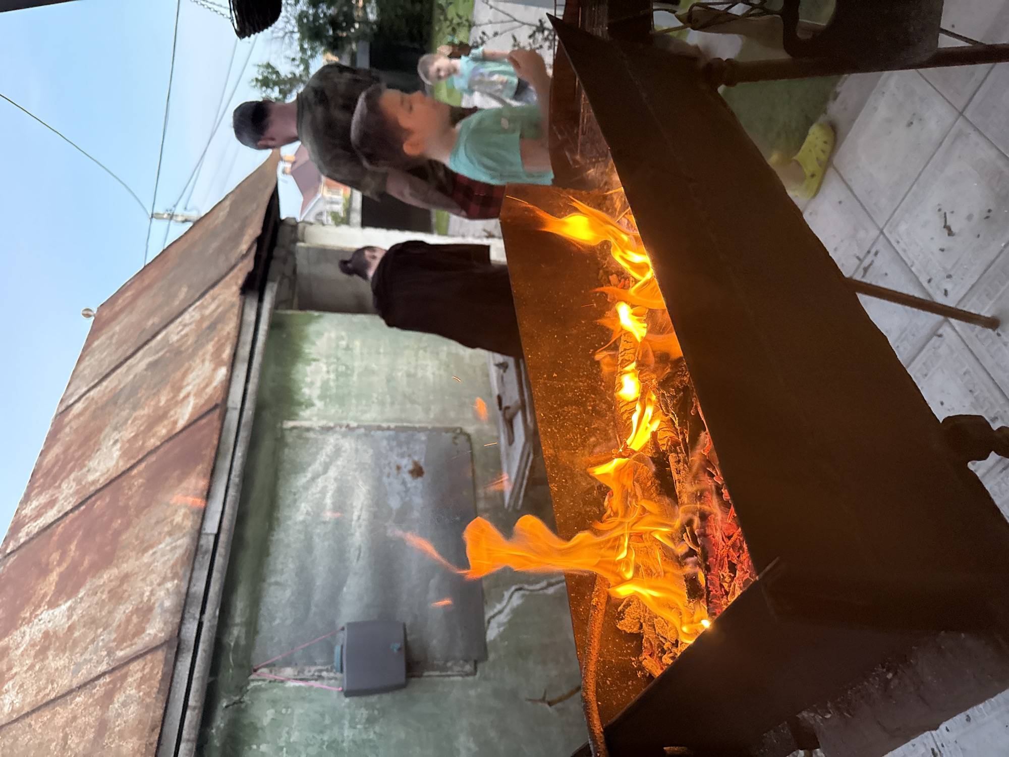This image, taken in Lipetsk, Russia, depicts an outdoor evening scene focused on a metal brazier (mangál) with an active wood fire. The brazier, angled from the lower left, contains vibrant orange and yellow flames rising from glowing red embers. Small sparks are visible above the fire.
In the foreground right, a young boy in a light green t-shirt and bright yellow Crocs stands, observing the fire with his right hand slightly raised. Behind him and to the right, an adult male, viewed from the back, wears a camouflage-patterned t-shirt and plaid shorts. To the mid-ground left, an adult female, also viewed from behind, wears a dark long-sleeved garment and has her hair styled in a bun; she is positioned near a building. Further in the background right, another child in a light blue t-shirt is partially visible.
The building in the mid-ground left has faded green/teal painted walls and a heavily rusted, corrugated metal roof. A dark gray rectangular container, possibly a water tank or electrical box, is mounted on the wall, secured by a pink strap. The ground in the immediate foreground consists of light-colored tiles with scattered dark specks. Beyond the tiled area, a grassy section is visible. The sky is a clear light blue, indicative of dusk. In the distant background, residential structures with reddish roofs and a utility pole with overhead wires are discernible. A dark hanging object is partially visible in the upper right corner.
