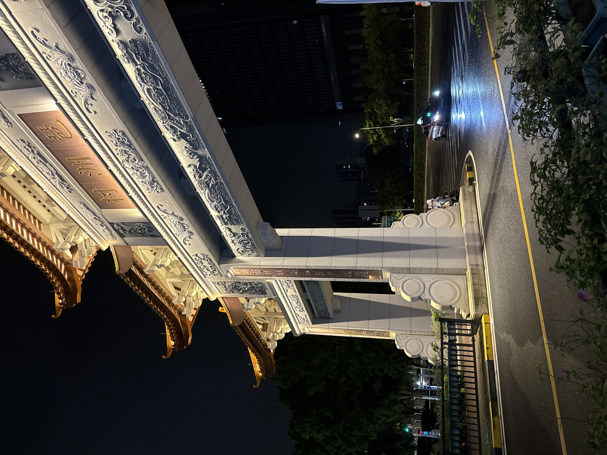 The image shows a large, ornate Chinese-style archway at night in Haizhu District, China. The archway is illuminated with warm yellow lights, highlighting its intricate carvings of dragons and clouds. Golden Chinese characters are prominently displayed on the arch. To the right of the arch, a modern, dark building stands tall. In the foreground, a wet asphalt road with yellow and black markings curves around the base of the arch. A red car with its headlights on is driving across a crosswalk, its lights reflecting on the wet surface. A person on a scooter is stopped to the left of the car. Trees and other buildings are visible in the background. The overall scene suggests a vibrant urban environment at night, with traditional architecture juxtaposed against modern development.