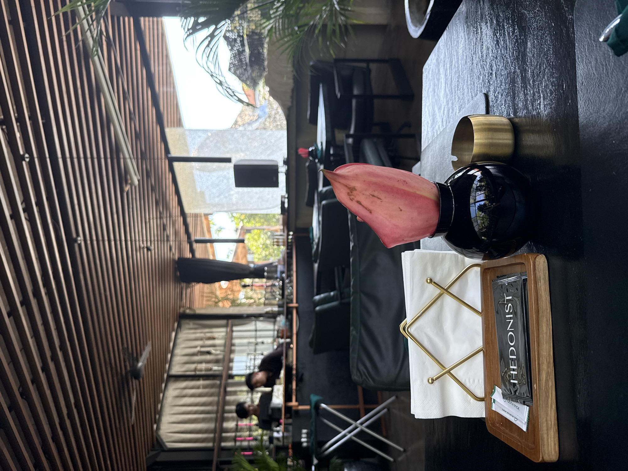 This image depicts a close-up view of a table setting at a restaurant or bar, likely located in Kuta Selatan, Indonesia, given the tropical flora visible in the background. The foreground features a dark, textured tabletop with a wooden tray holding a packet labeled "HEDONIST" and two smaller packets. A gold napkin holder with white napkins is positioned behind the tray. To the right of the napkins is a black, glossy vase containing a single, large pink petal. Adjacent to the vase is a small, brass-colored ashtray.
The mid-ground reveals seating areas with dark, possibly leather, chairs and tables. In the background, a bar area is visible with two individuals, appearing to be staff members, behind it. The bar is stocked with bottles and glassware. The overall ambiance suggests an indoor-outdoor setting, with open spaces and natural light filtering in. The ceiling is constructed with wooden slats, and a ceiling fan is visible. The decor hints at a contemporary and stylish establishment.