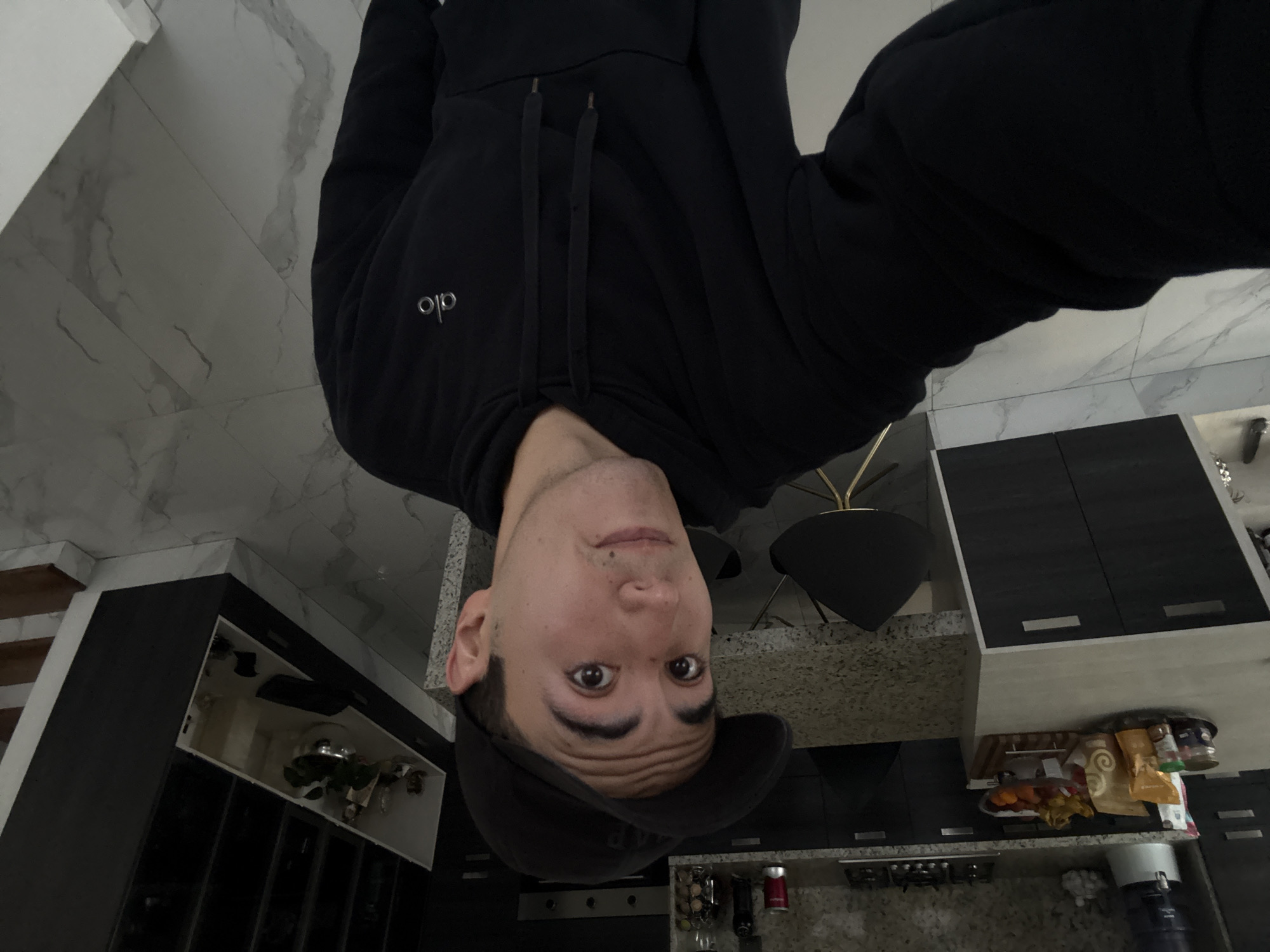 An inverted full-shot selfie captures a young man standing upside down in what appears to be a modern kitchen or dining area. The man, likely in his 20s or 30s, has short dark hair, dark eyes, and a neutral to slightly playful expression as he looks directly at the camera. He is wearing a black hoodie with drawstrings, featuring the "alo" logo in white on the left chest, and a dark baseball cap.
The room, also inverted, showcases a contemporary design. The floor (appearing as the ceiling in this orientation) is a light grey with a marble-like pattern. The background features dark cabinetry, some with white or light gray accents, suggesting kitchen cupboards or storage units. One cabinet on the left is open, revealing various items inside, possibly dishes or small decorative plants. Another section of cabinetry on the right shows items on the counter below, including a bowl of fruit, several jars, bottles, and what looks like a coffee maker. A dark chair with a golden base is partially visible on the left side of the frame. The countertops have a speckled, granite-like texture.
The overall impression is a casual, spontaneous selfie taken indoors. The time of day is not discernible from the lighting. The location context provided is San Bernardino Tlaxcalancingo, Mexico, suggesting this is a home environment.