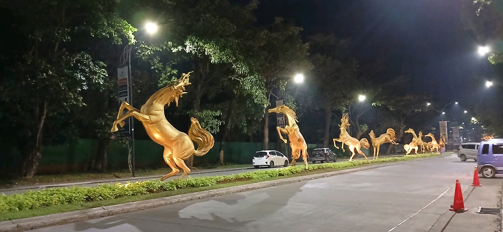 The image depicts a nighttime street scene in Cirebon, Indonesia, featuring a row of large, golden horse sculptures lining a median strip. The sculptures appear to be in various dynamic poses, some rearing up. The street is paved and appears to be a main thoroughfare with traffic lights and street lamps visible. Several vehicles are present, including a white hatchback, a dark SUV, and a purple van. Red traffic cones are placed on the side of the road. Lush green vegetation forms the median strip, and mature trees with dense foliage line the street, illuminated by the artificial lights. The overall impression is that of a decorative street enhancement, possibly for a festival or public art installation.
