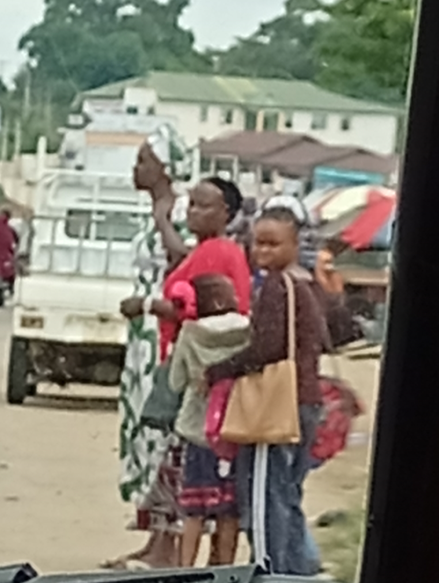 A group of people, including women and children, are standing on a dirt road. The main subjects are three women and a child. The woman on the left is wearing a green and white patterned dress and a headscarf. The woman in the middle is wearing a red top and is holding a child who is wearing a grey hoodie and shorts. The woman on the right is wearing a brown sweater and jeans and carrying a tan bag. They appear to be waiting or walking, possibly near a market or roadside stalls, as evidenced by the awnings and a white truck in the background. The environment suggests a casual outdoor setting in a populated area.