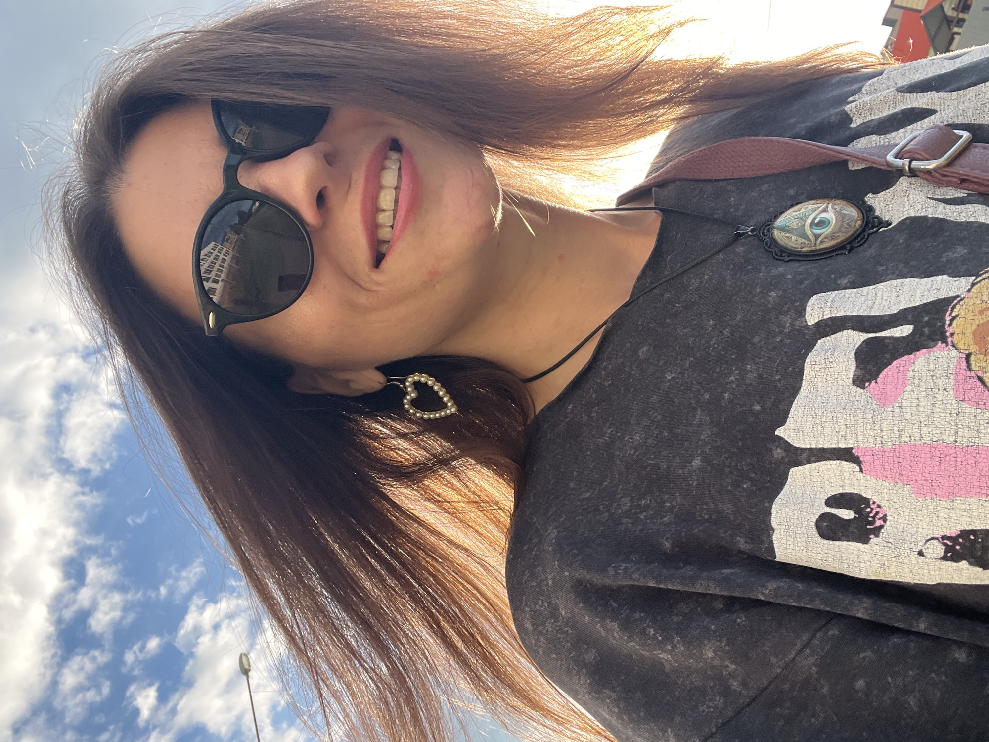 This is a close-up, low-angle selfie of a young woman in Krasnoe-na-Volge, Russia. She is outdoors, with a bright blue sky and scattered white clouds filling the background, suggesting it's daytime, likely late afternoon due to the warm, golden sunlight backlighting her hair and face. She is wearing round, black sunglasses that reflect a building, and her mouth is open in a wide, joyful smile, showing her teeth. Her long, brown hair is windswept and catches the light, adding to the sense of movement and a carefree mood.
She is wearing a dark grey t-shirt with a faded, possibly graphic print. Around her neck, she wears a necklace with a pendant that resembles an eye within an ornate, dark frame. A brown strap, possibly from a bag or backpack, crosses her shoulder. In her hair, near her ear, a small heart-shaped accessory adorned with pearls is visible. The overall impression is one of happiness and enjoying a sunny day outdoors. There is no visible text in the image.