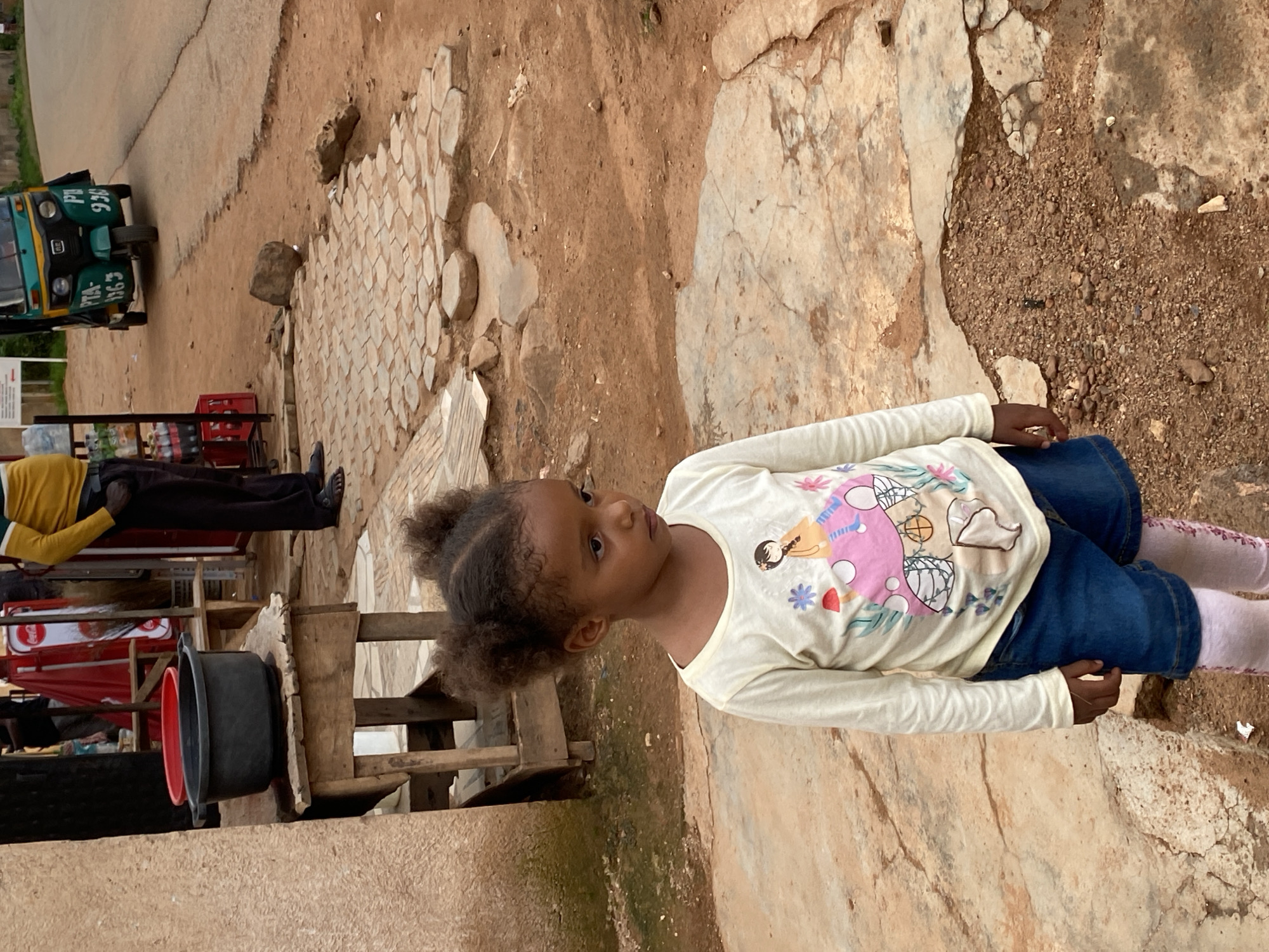 A young girl stands on a dirt and cracked concrete ground in Gura Top, Nigeria. She has dark, curly hair styled in two small buns. She is wearing a long-sleeved cream shirt with a cartoon illustration and blue denim shorts, over pink patterned tights. Behind her, a wooden table holds a black basin and a red bowl. In the background, a person in a yellow and dark striped shirt and dark pants stands near a stall with Coca-Cola branding. A green auto-rickshaw is parked on the road to the right. The environment appears to be a rural or semi-urban area with red dirt and scattered stones.