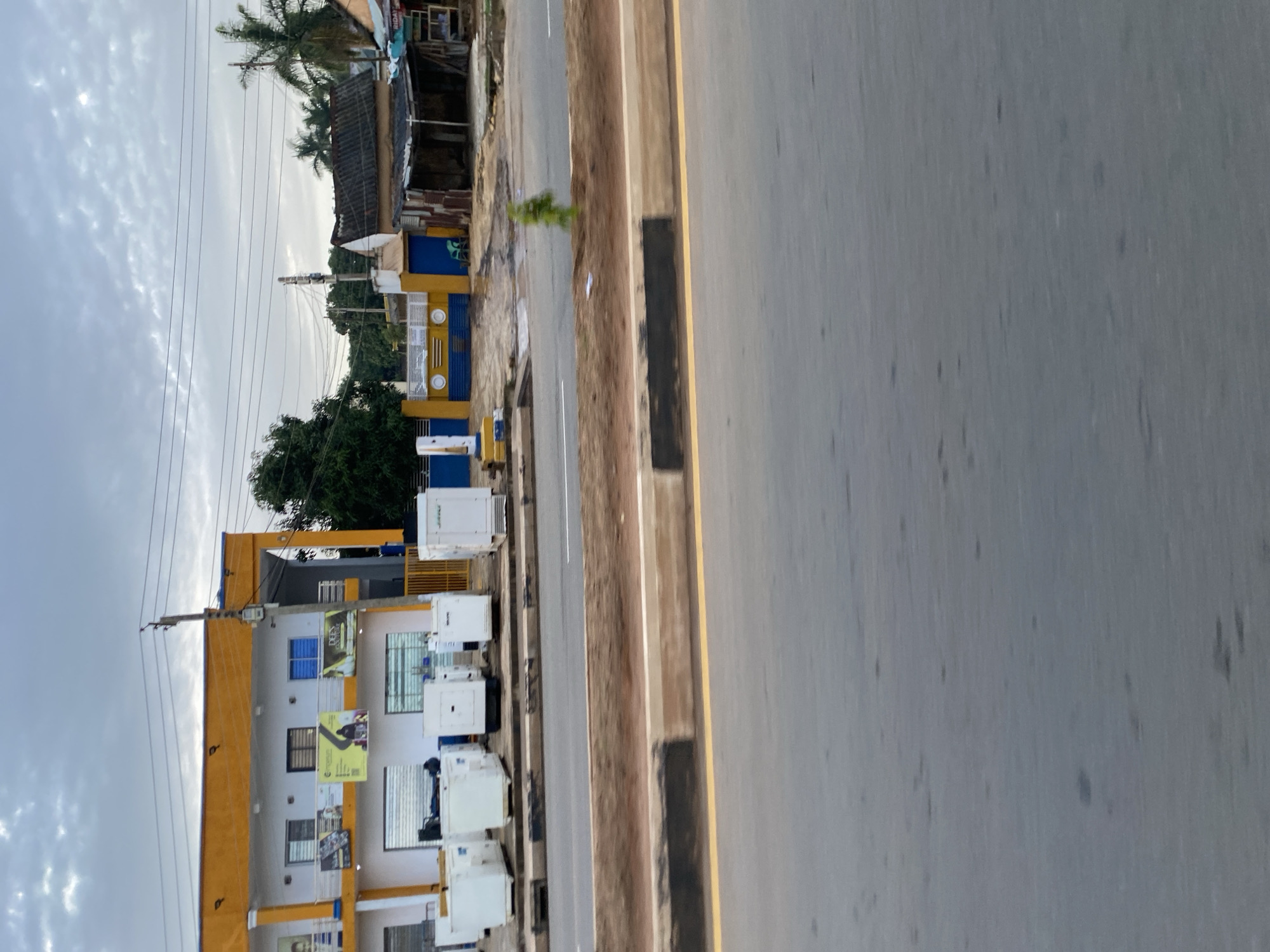 This image depicts a street scene in Jos, Nigeria, featuring a commercial building with an orange and white facade. In front of the building, several white generators are lined up along the sidewalk. The building has multiple windows and balconies, with signage visible on the upper floors. Adjacent to the commercial building, there is a yellow and blue fence enclosing a smaller property with a dilapidated structure made of corrugated metal. The street in the foreground is paved, with a clear yellow line marking the lane. The sky is overcast with scattered clouds. There are no people clearly visible or actively engaged in any interaction in the immediate foreground or on the street. The overall environment suggests a commercial area, likely in a developing urban setting.