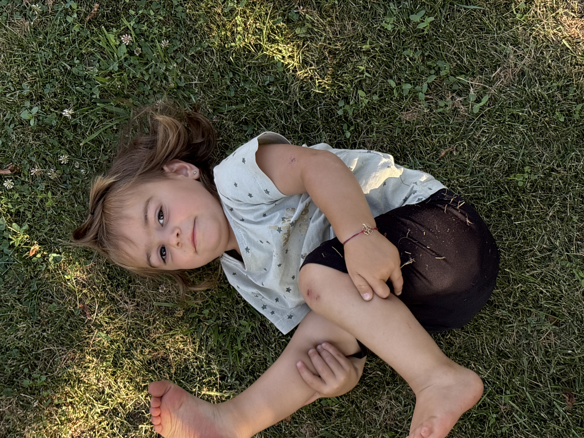 A young girl with light brown hair lies on her back in a grassy area. She is wearing a light blue t-shirt with small white stars and black shorts. Her right arm is bent at the elbow and her hand is resting on her thigh. A thin red bracelet with a four-petal flower charm is visible on her wrist. She has a small abrasion on her right knee and elbow. Her left leg is bent at the knee, and her left foot is visible in the lower left corner of the frame. The girl is looking up and to her left, with a slight smile on her face.
The scene appears to be outdoors in a park or yard, with dappled sunlight suggesting it is daytime, likely late afternoon due to the angle of the light. The grass is green and appears healthy, with a few small white flowers scattered throughout. The setting is likely Oreshak, Bulgaria. There is no visible text in the image. The overall mood is relaxed and carefree.